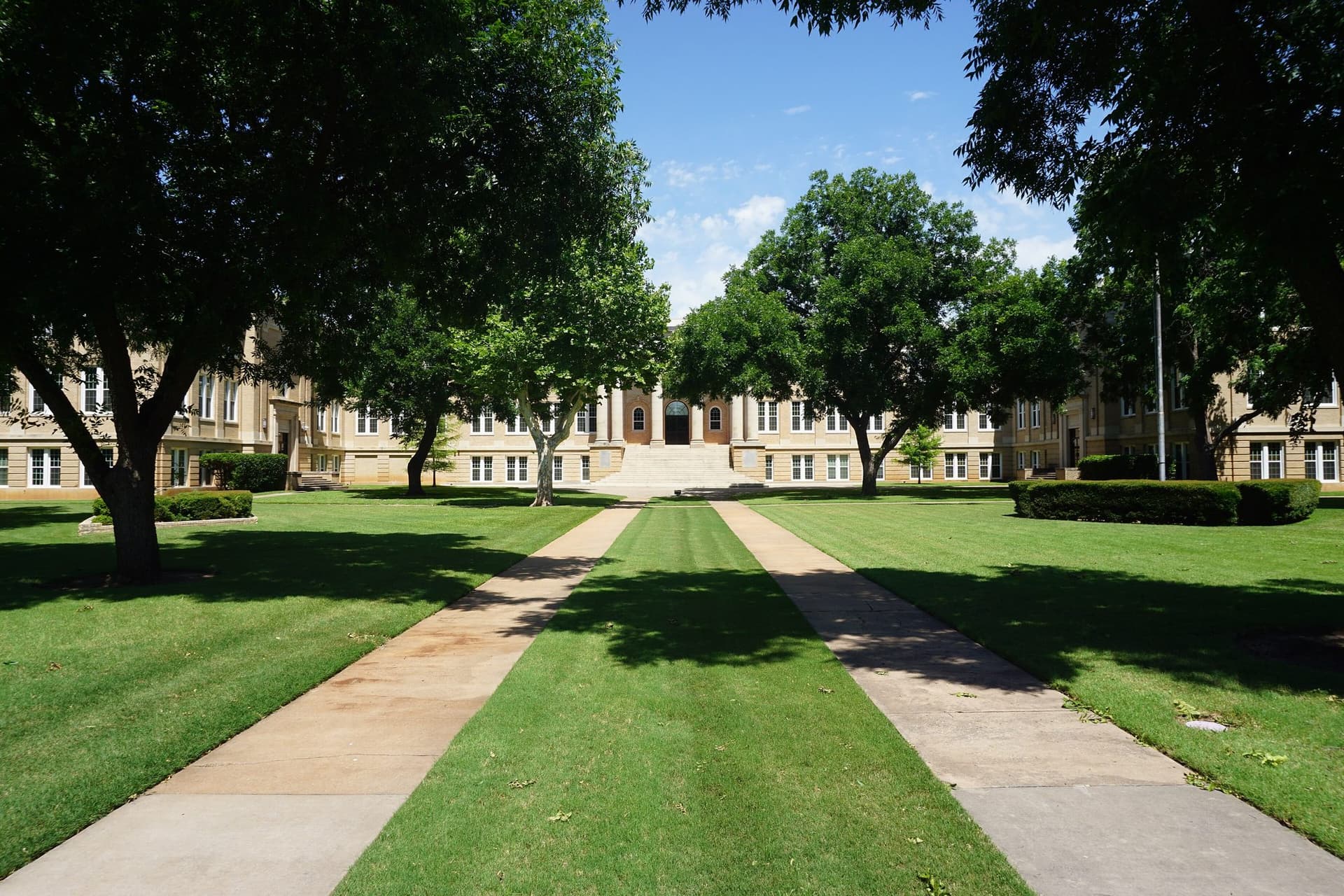 A tan brick, three-story administration building with a columned central entrance and wide steps, seen down a long lawn between two concrete walkways shaded by large green trees under a blue summer sky