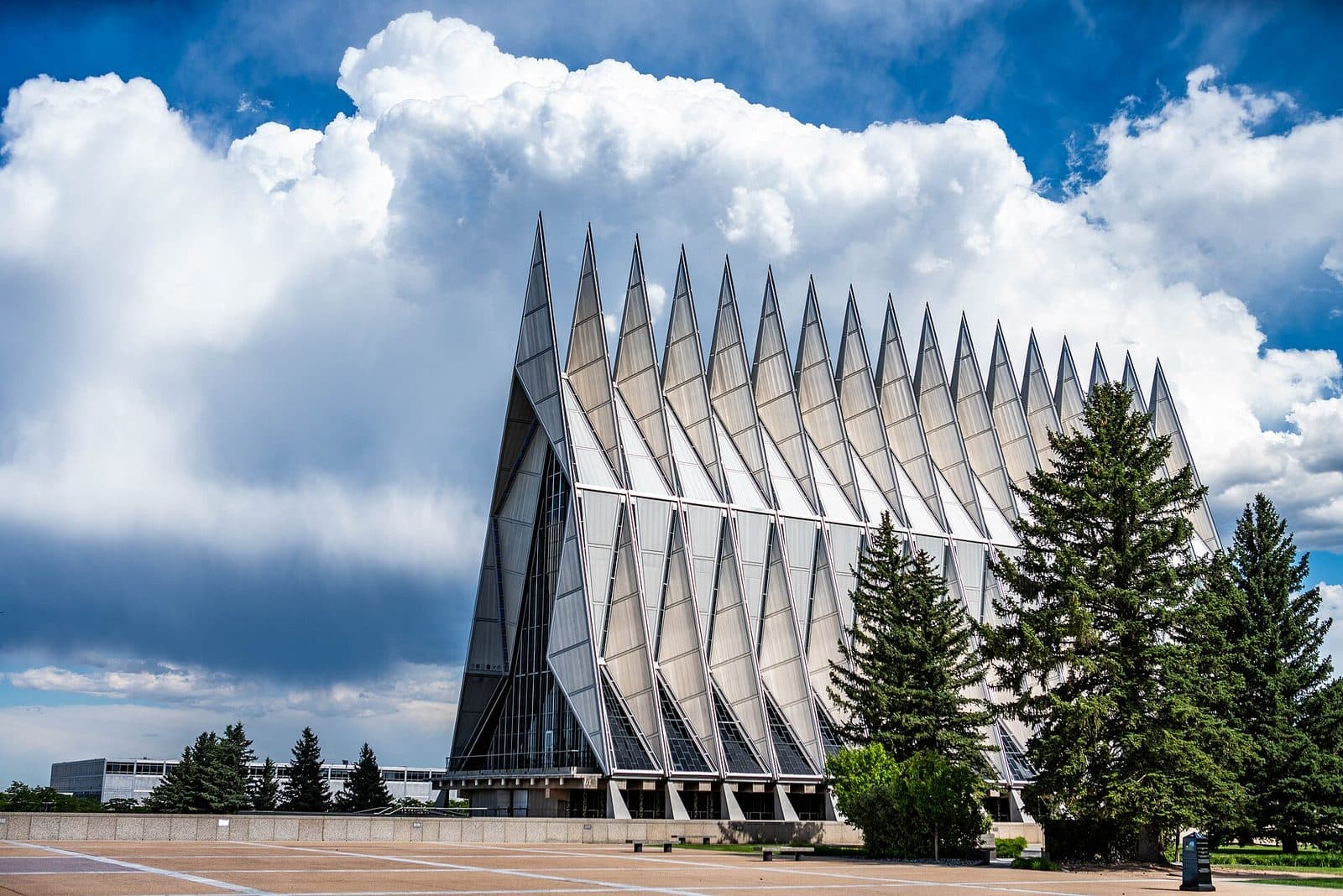 The aluminum spires of the Air Force Academy Cadet Chapel against a blue sky with large white clouds, with evergreen trees at right and a paved plaza in front
