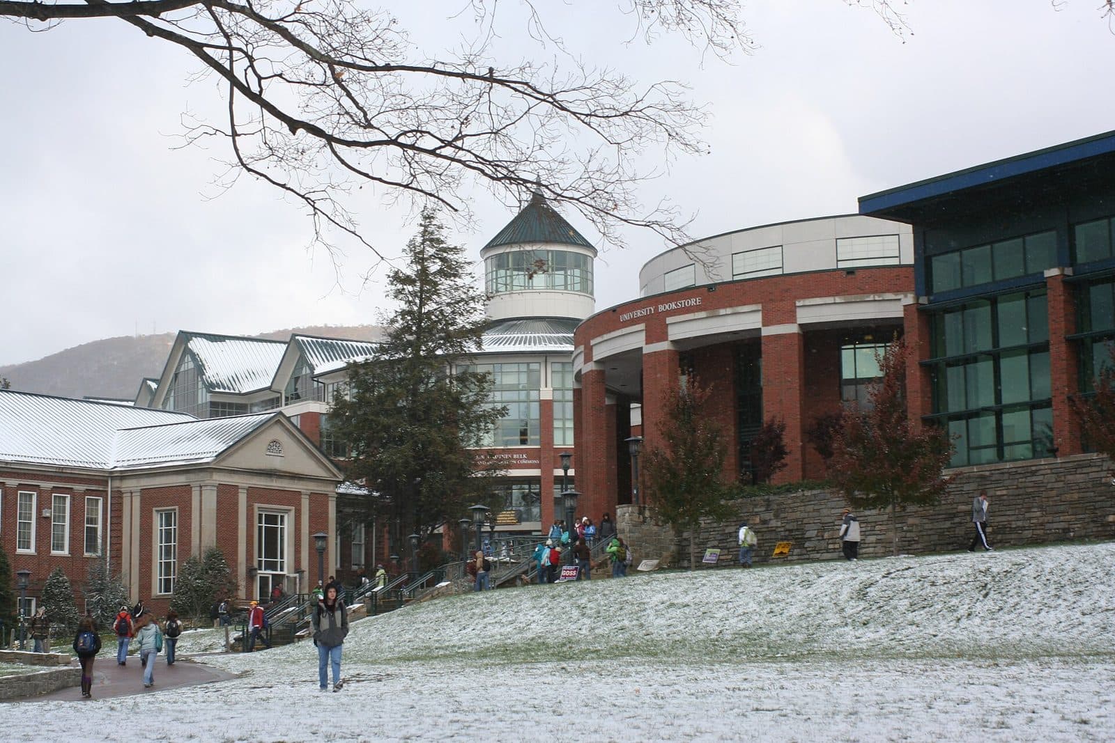 A light snow on Sanford Mall at Appalachian State, with students walking toward the brick library and student union buildings and a mountain ridge behind