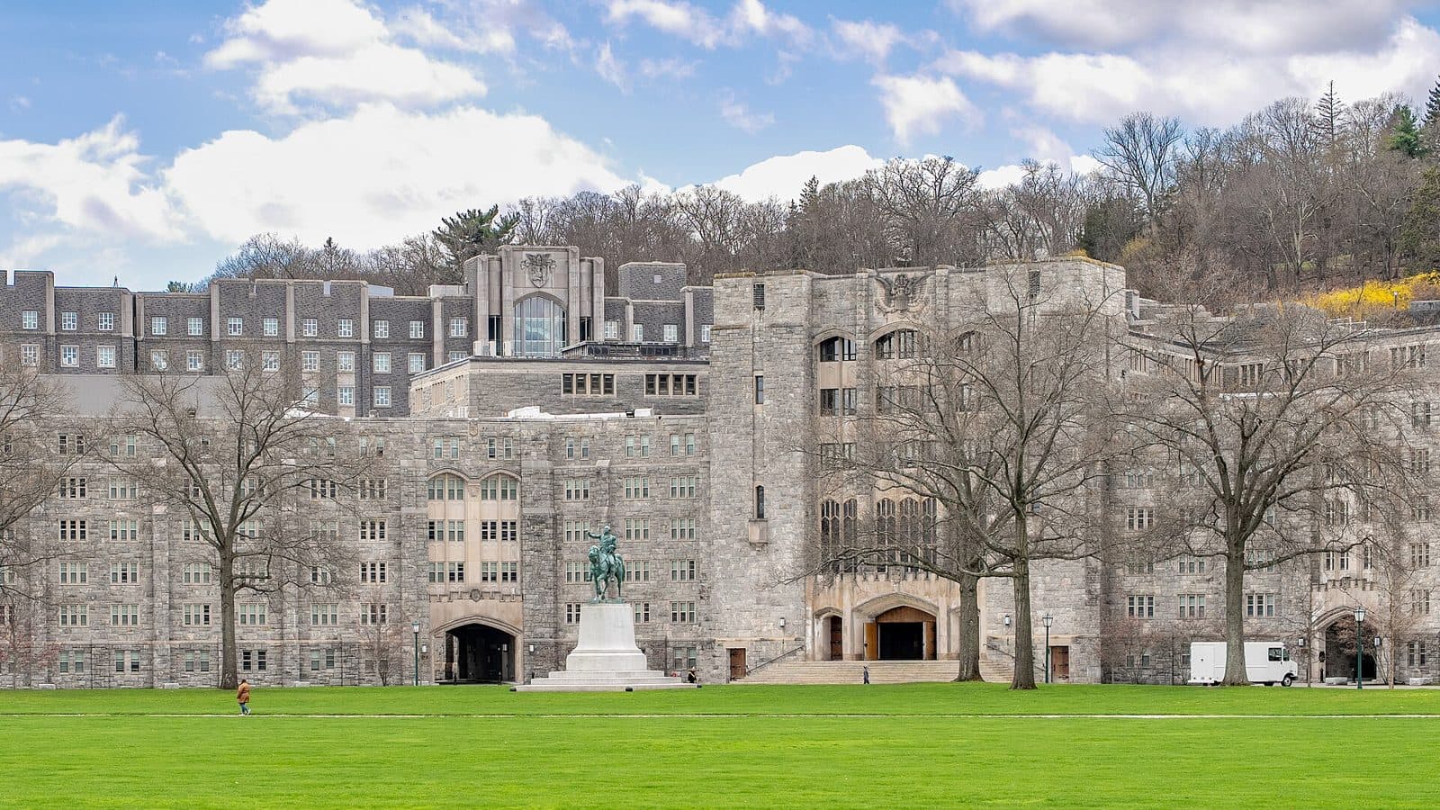 Gray granite gothic-style buildings behind a wide green lawn, with an equestrian statue on a white pedestal, bare trees and a wooded hill under a partly cloudy blue sky