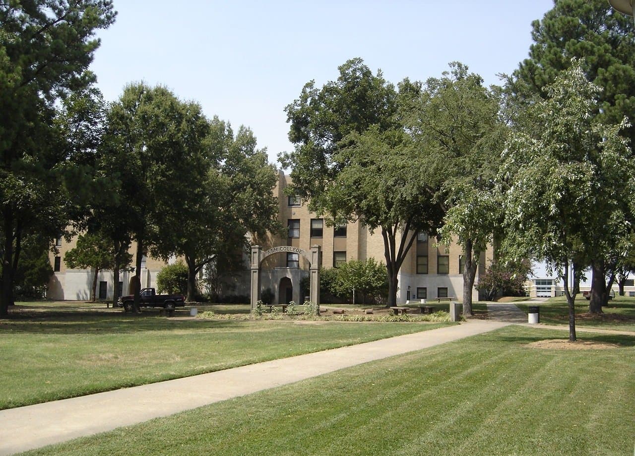 A tan brick three-story campus building behind a stone archway, seen across a mowed lawn with a curving sidewalk and tall shade trees on a clear summer day