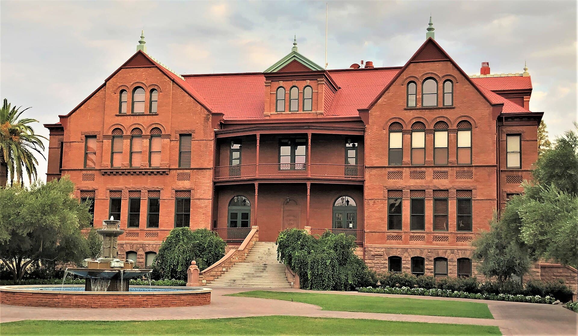 Old Main at Arizona State University, a three-story red-brick Victorian building with a red roof, gables and a central balcony, behind a tiered fountain, lawn and a palm tree