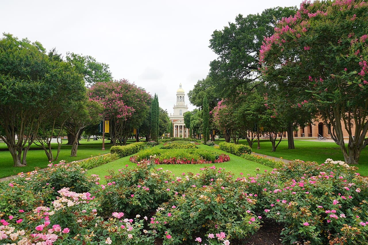 A wide brick-paved mall lined with trees leading to Pat Neff Hall, a red-brick building with white columns and a tall domed cupola, under a blue summer sky