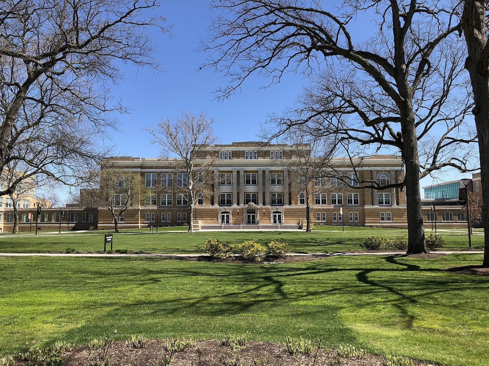 The tan brick and stone facade of University Hall at Bowling Green State University, seen across a green lawn framed by bare spring trees