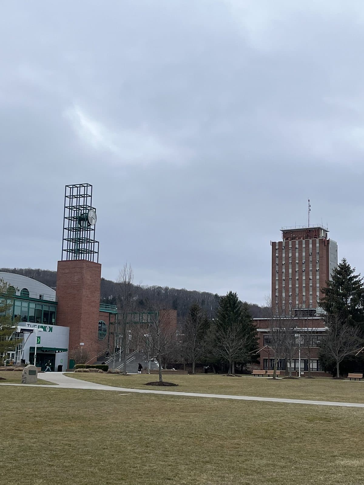 A brick clock tower topped with an open steel frame beside the glass-fronted University Union, with a tall brick tower and wooded hills behind a wide lawn at Binghamton University