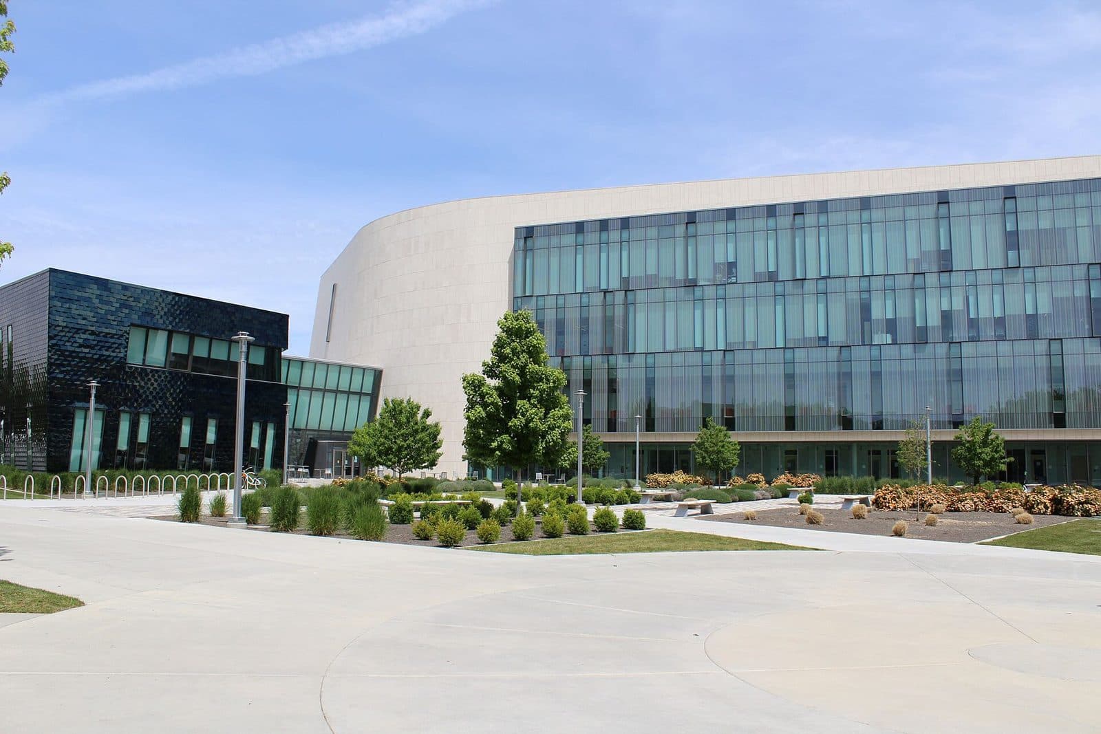 A modern curved building with a pale stone wall and rows of blue-green glass windows, beside a smaller dark-tiled glass building, behind a wide concrete plaza with young trees and landscaped beds under a blue sky