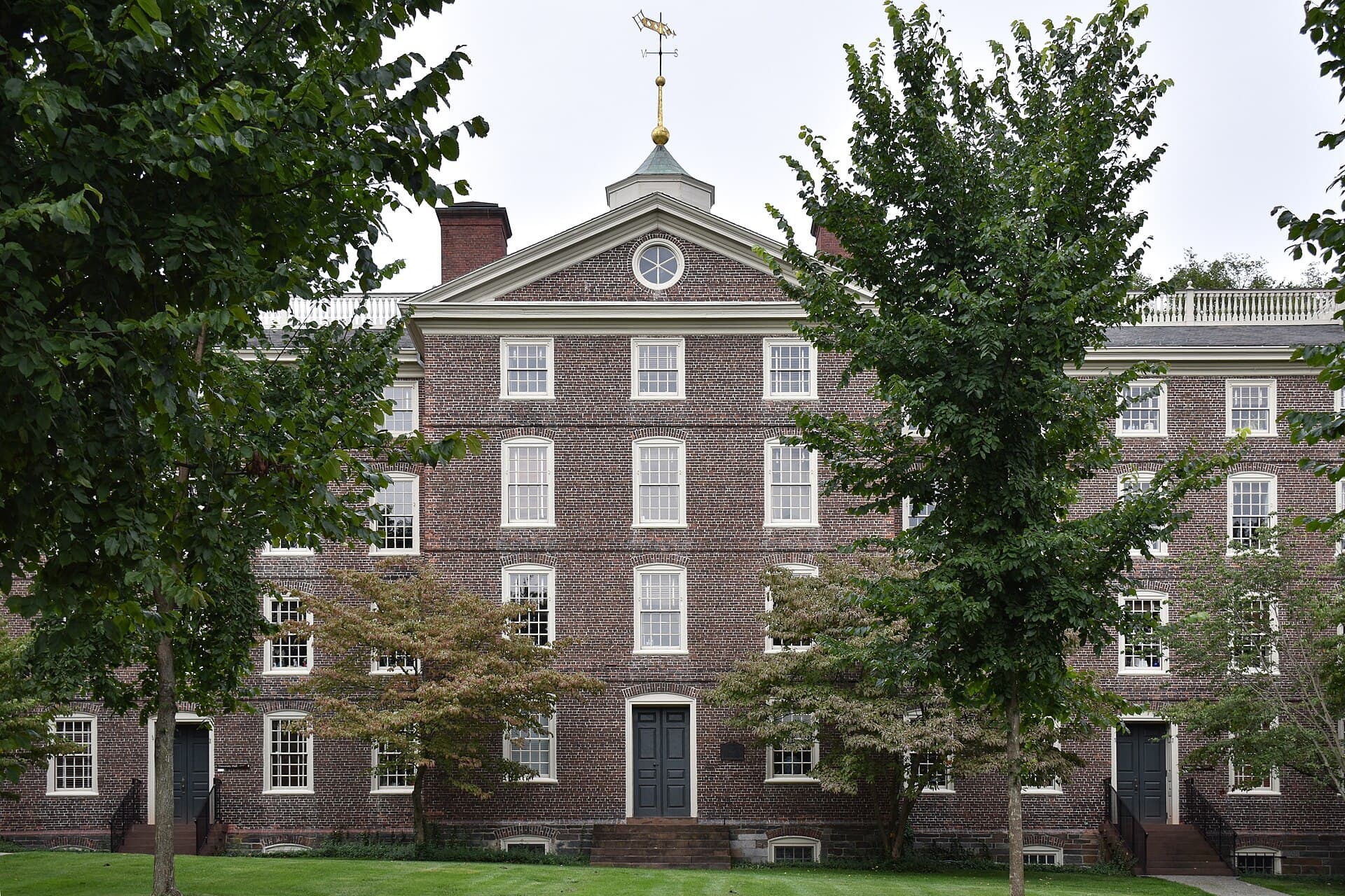 University Hall at Brown, a four-story brick building with a white cupola and weathervane, behind leafy trees on the Main Green