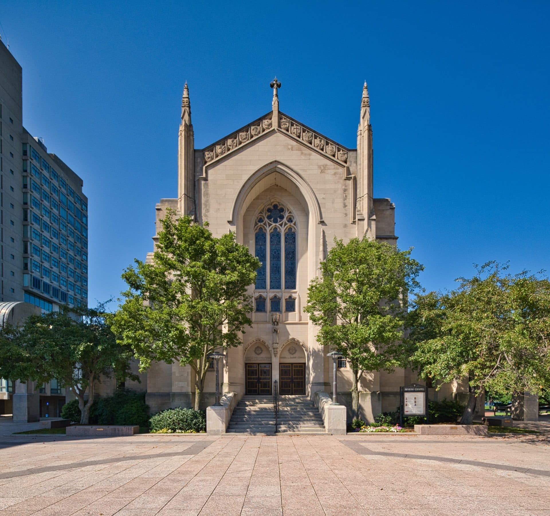 The Gothic stone facade of Marsh Chapel at Boston University, framed by leafy trees on Marsh Plaza under a deep blue sky