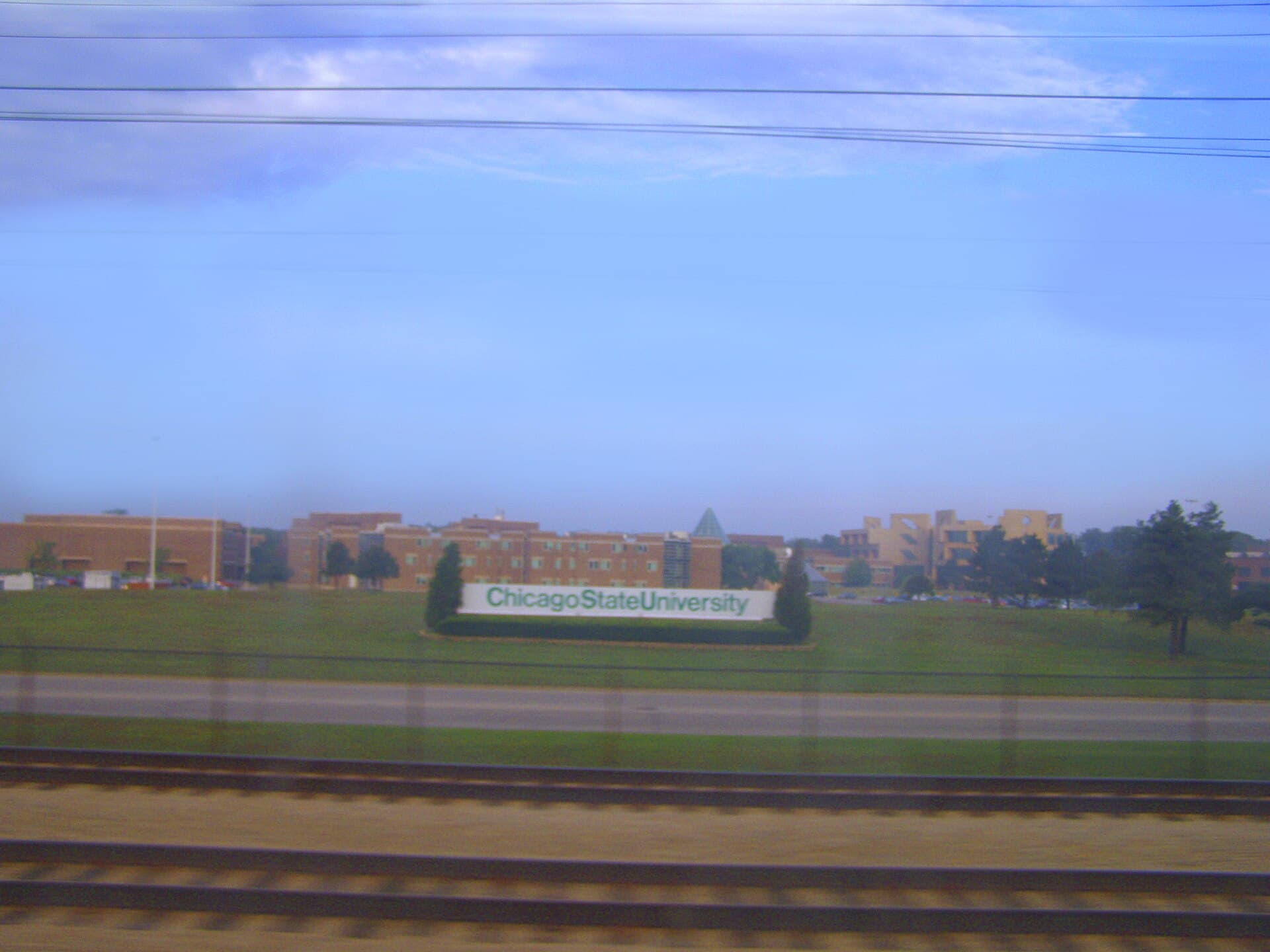 A green-and-white Chicago State University sign on a wide lawn in front of low red-brick and tan campus buildings, seen past railroad tracks under a pale blue sky