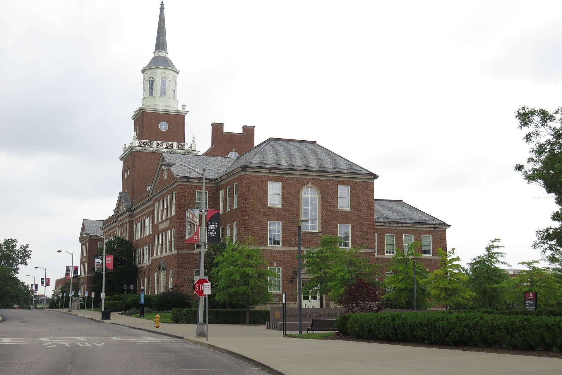 The red-brick McMicken Hall at the University of Cincinnati, with its white clock tower and cupola, behind a lawn and hedges along a campus road