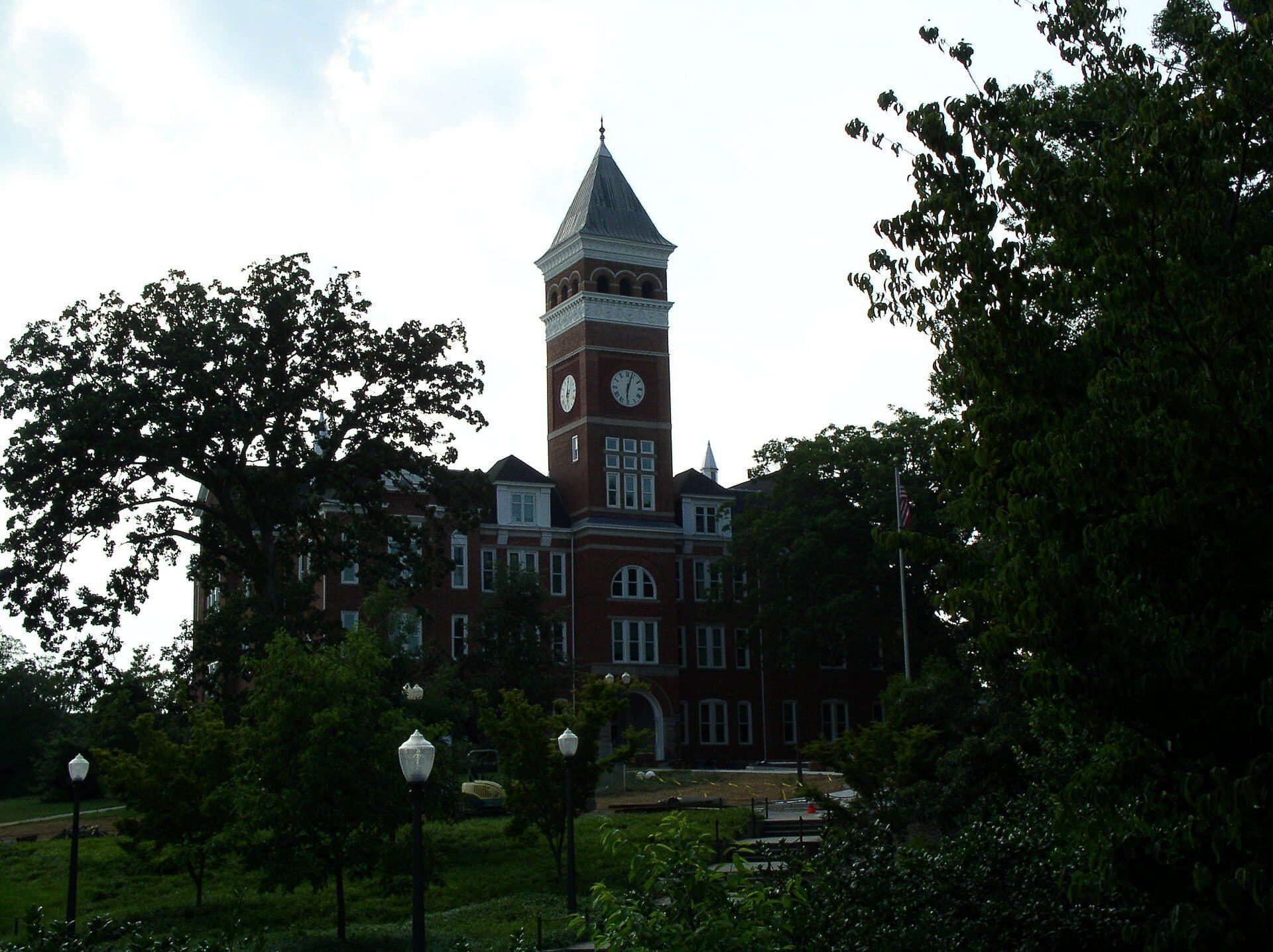 Tillman Hall's red-brick clock tower rising above trees and a lawn with lampposts at Clemson University