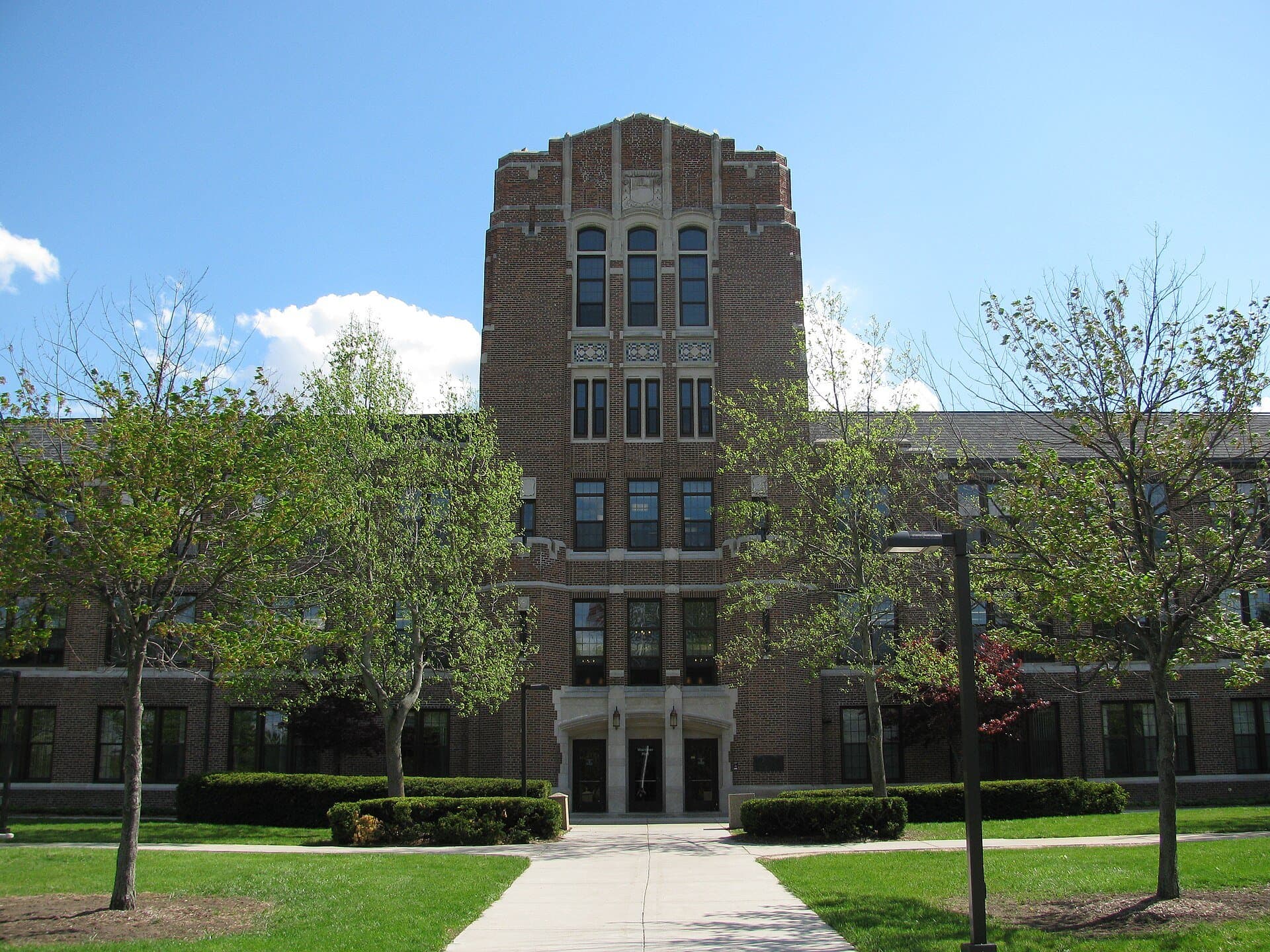Warriner Hall at Central Michigan University, a brown-brick building with a tall central tower, behind a long walkway lined with young trees and lawn