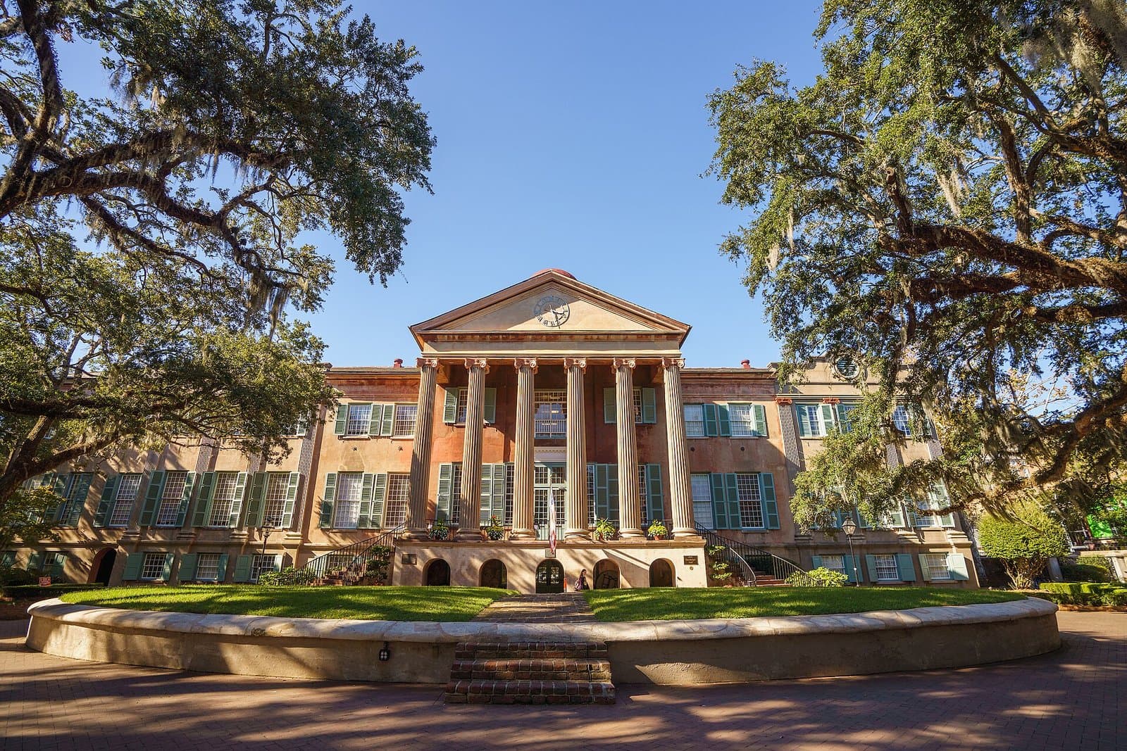A three-story peach-colored building with green shutters and a tall white-columned portico, seen across a round raised lawn and brick paving, framed by live oaks hung with Spanish moss under a deep blue sky