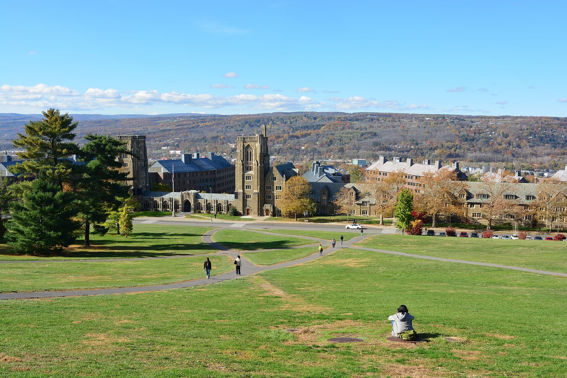 Looking down Cornell's grassy Libe Slope toward stone campus buildings and a clock tower, with wooded hills beyond