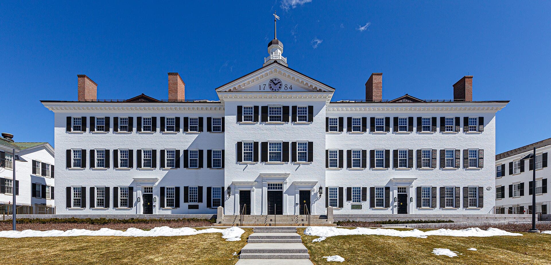 White Dartmouth Hall with its cupola and rows of dark-shuttered windows, under a deep blue sky with patches of snow on the lawn