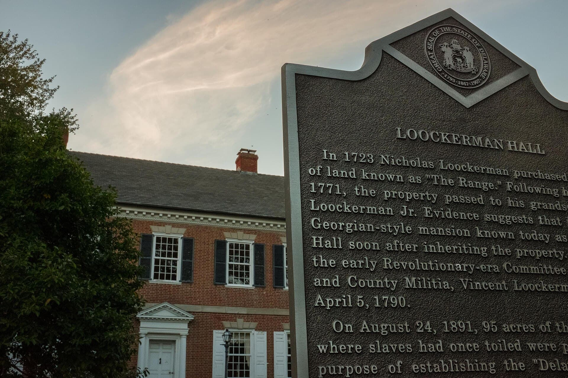 A red-brick Georgian mansion with black shutters, white-trimmed windows and a white doorway beside a tall gray historical marker titled Loockerman Hall, framed by a leafy tree under a dusky sky with a wisp of cloud