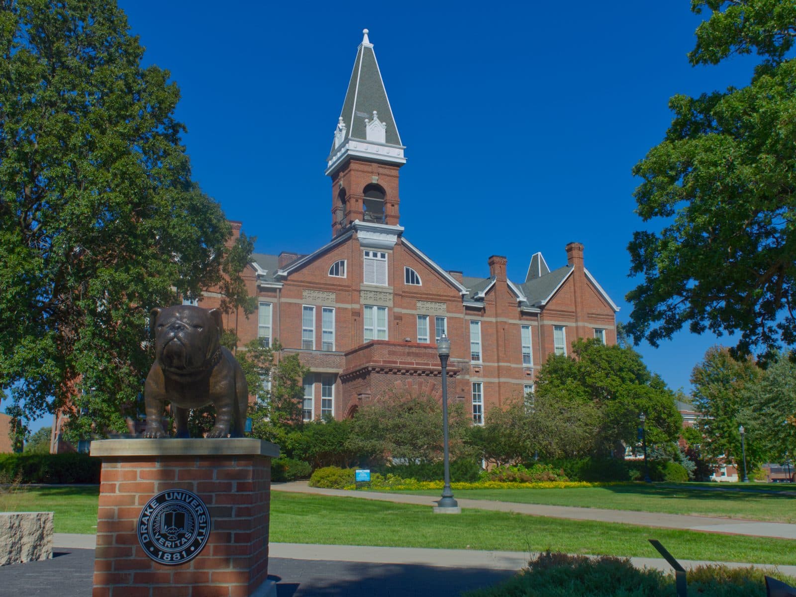 A bronze bulldog statue on a brick pedestal with the Drake University seal in front of Old Main, a red-brick building with a tall spire, under a deep blue sky