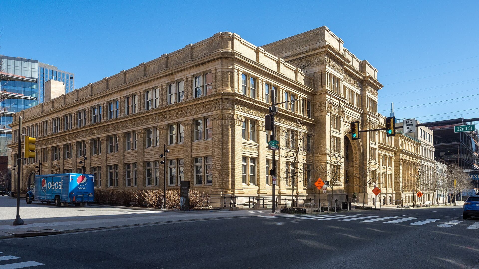 Drexel University's Main Building, a large ornate tan stone and brick building on a Philadelphia street corner, with traffic lights and a 32nd Street sign under a blue sky