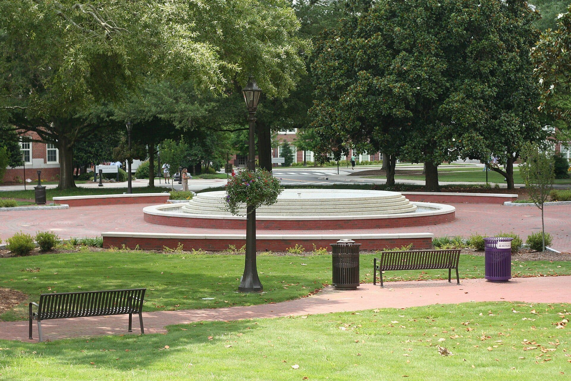 A round tiered fountain on a brick plaza in East Carolina University's tree-shaded quad, with lamp posts, benches and red-brick buildings behind