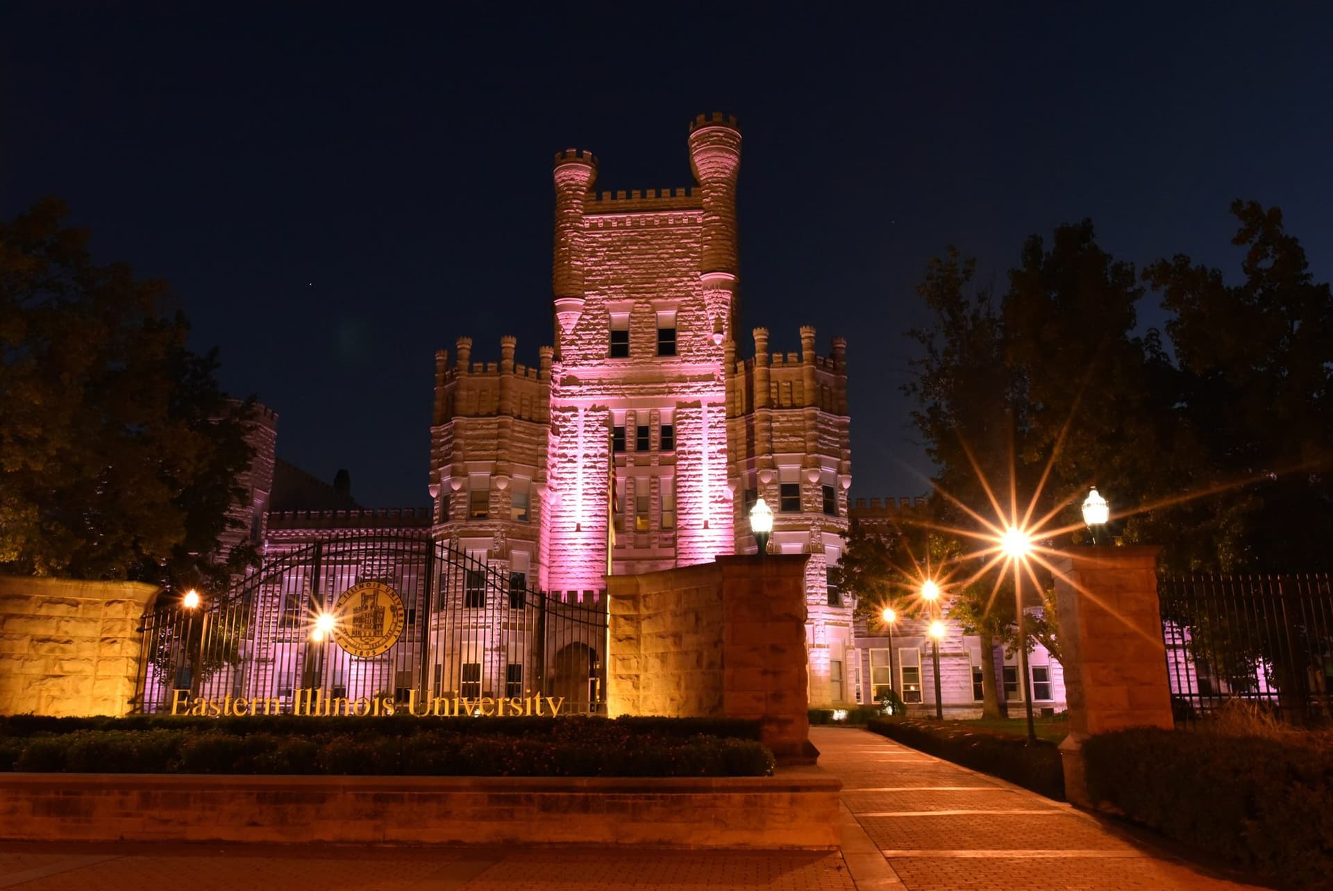 The castle-like stone Old Main building with crenellated towers lit in pink at night, behind an iron gate and stone wall with the words Eastern Illinois University in gold letters, and street lamps glowing along a brick walkway