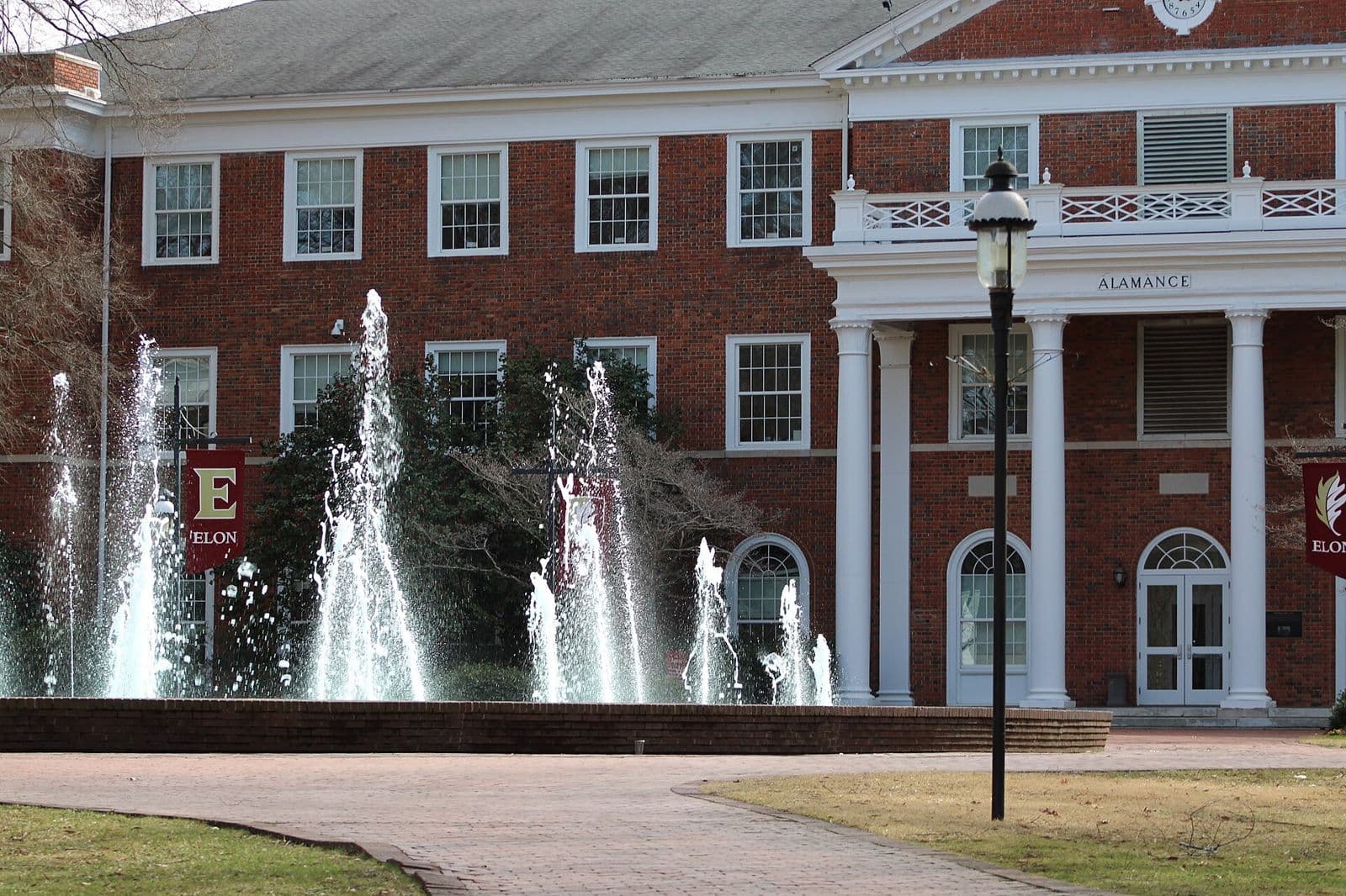 A red-brick building with white columns and a lamp post at Elon University, with a fountain spraying in the foreground