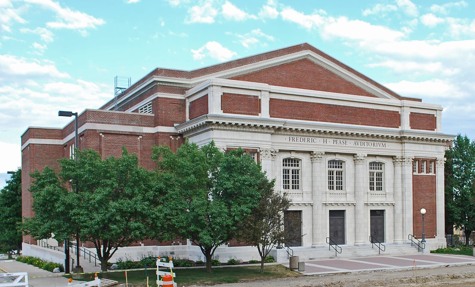 Pease Auditorium at Eastern Michigan University, a red-brick building with a white stone columned front and a triangular gable, behind green trees under a blue sky