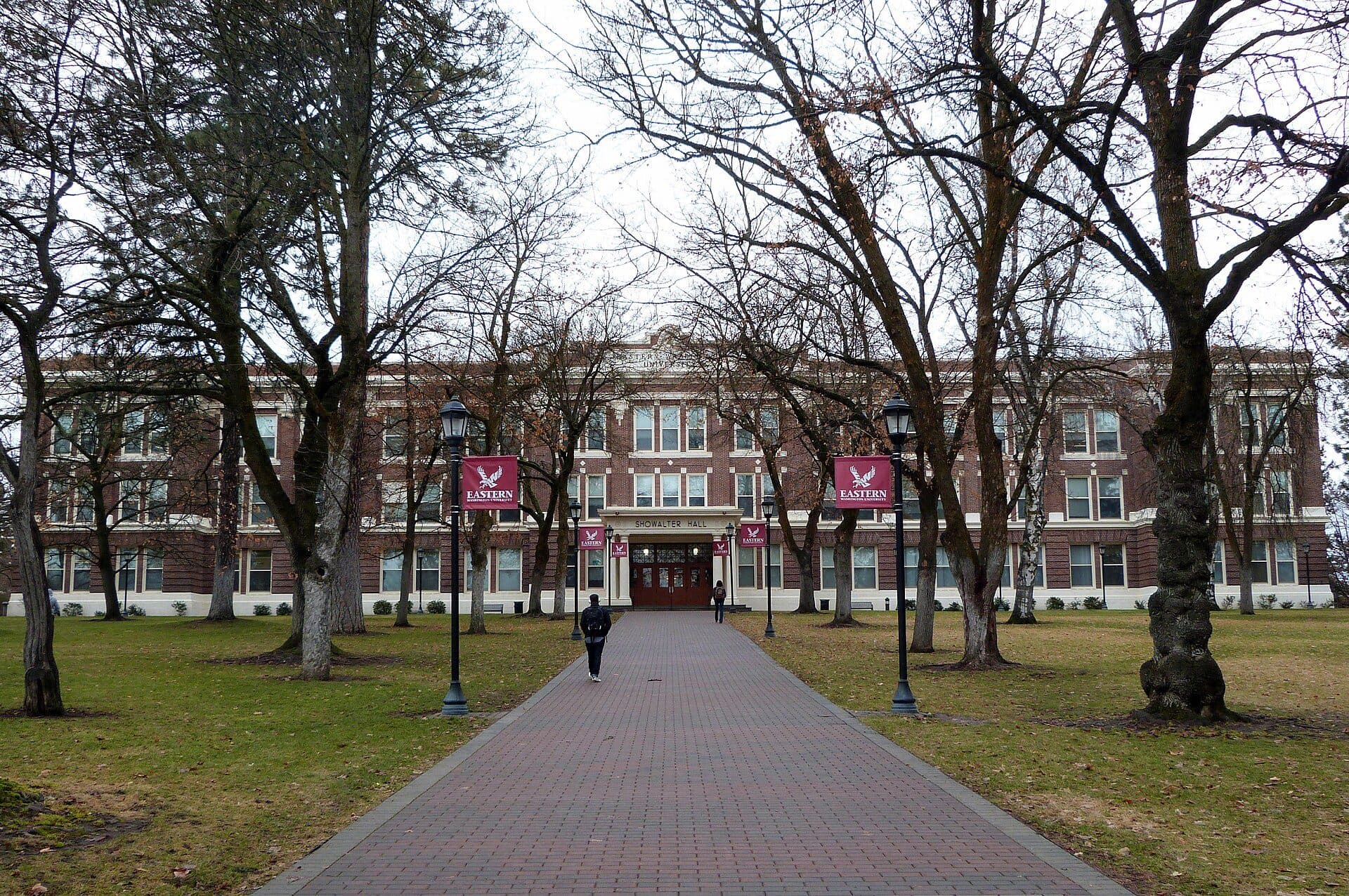 A brick walkway lined with bare trees and red Eastern banners leading to Showalter Hall, a long red-brick building in Cheney