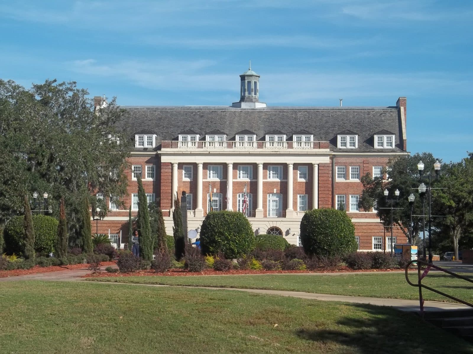 A three-story red-brick hall with a white-columned portico, dormer windows and a cupola on a slate roof, behind rounded green shrubs, a curving path and a sunny lawn under a blue sky