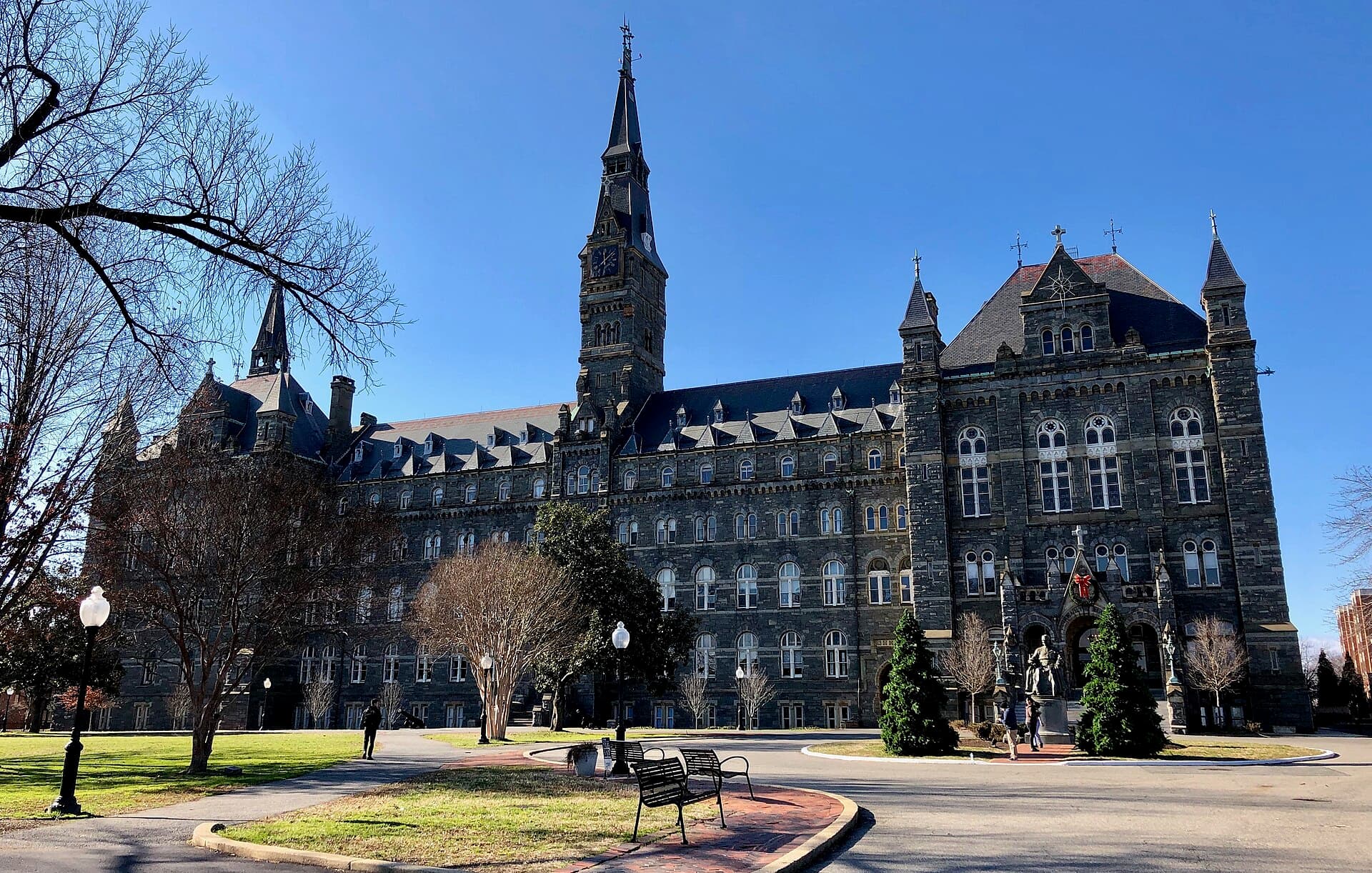 Healy Hall at Georgetown, a dark gray stone Gothic building with a tall clock tower and spires, behind a lawn with bare trees, benches and two small evergreens under a clear blue sky