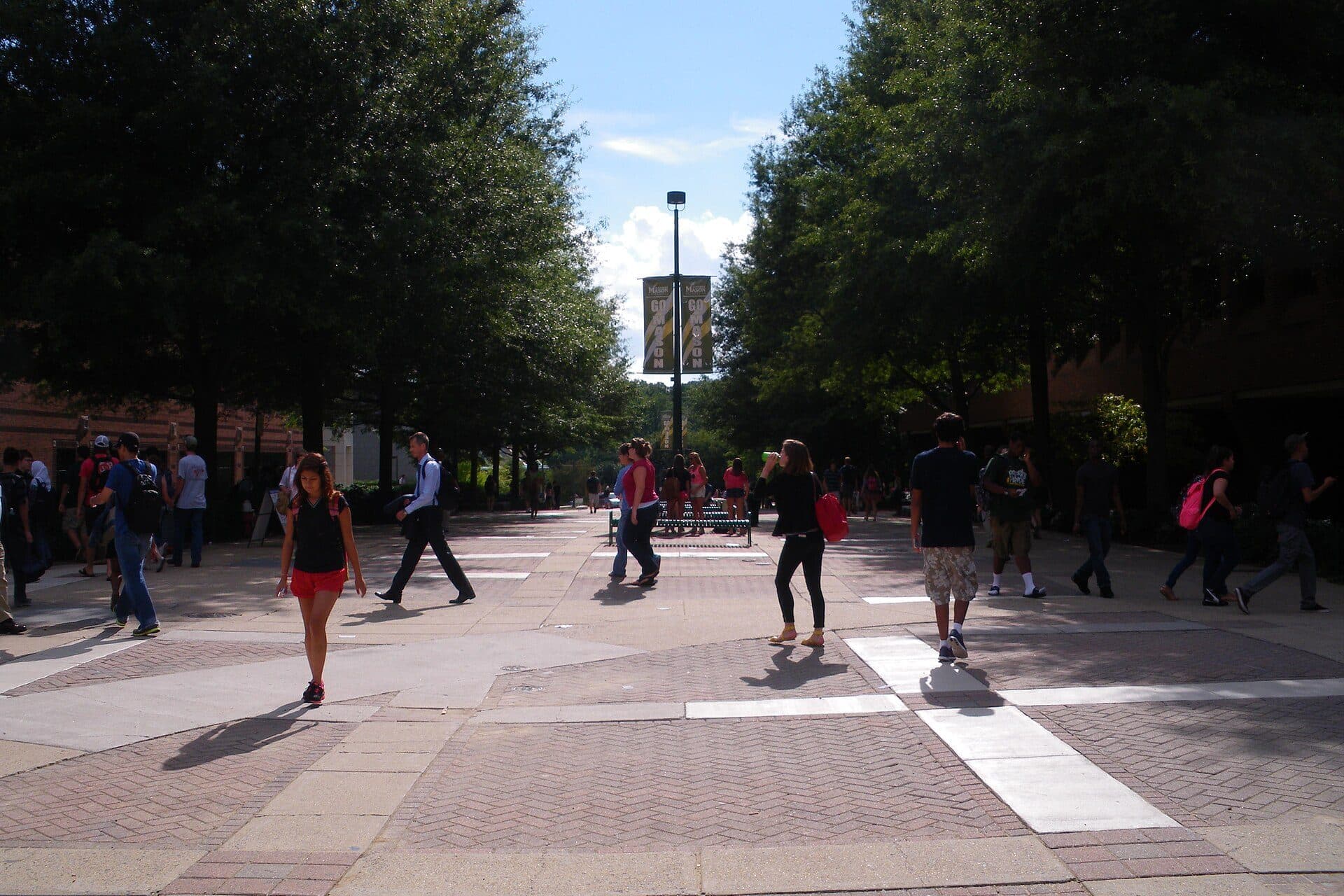 Students walking along a wide brick plaza lined with tall trees on George Mason's Fairfax campus