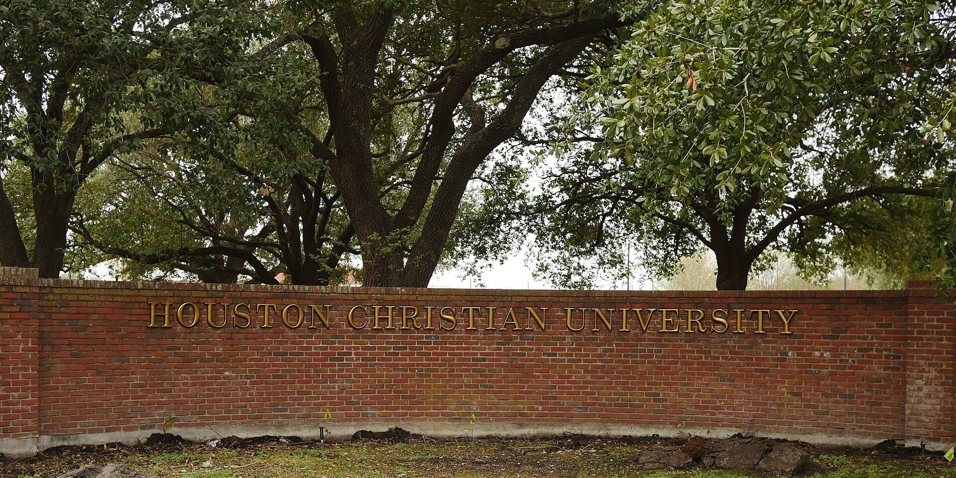 A long red-brick wall with brass lettering reading Houston Christian University, beneath the spreading branches of large live oaks
