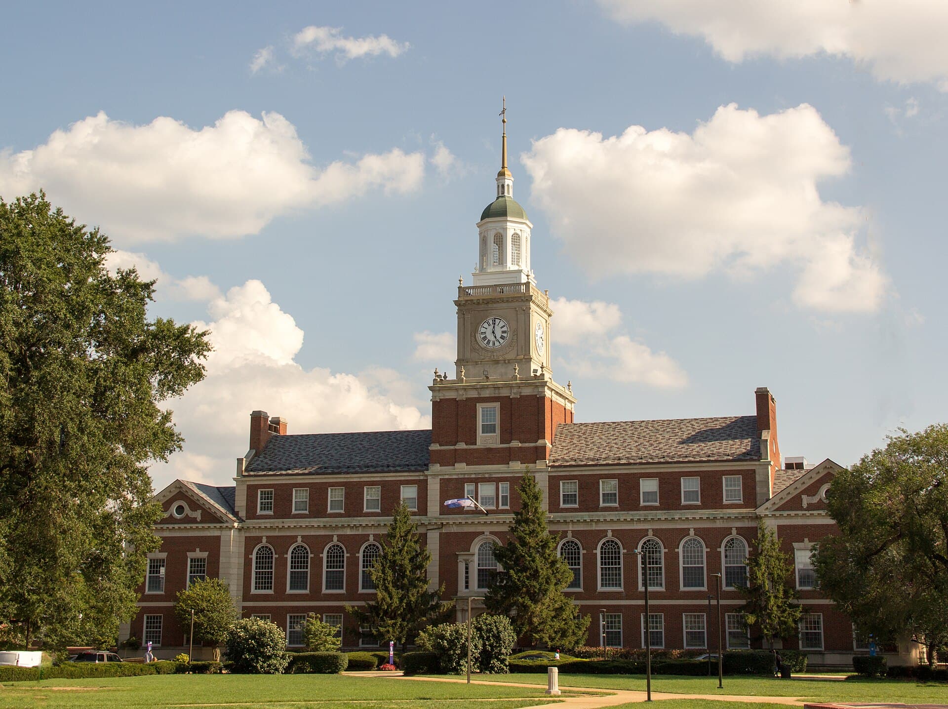 Howard University's Founders Library, a red brick Georgian building with a white clock tower and green cupola, behind a wide lawn and tall trees under a blue sky with white clouds