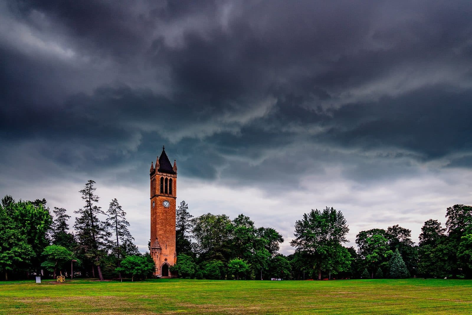 Iowa State's red-brick Campanile clock tower rising above a green lawn and tall trees under dark, stormy clouds