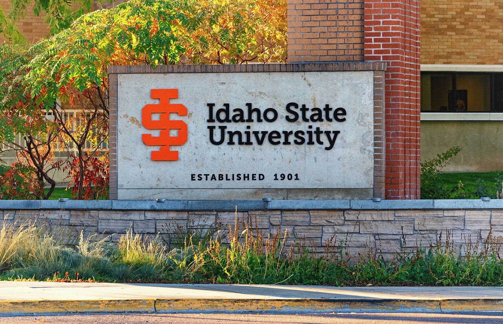 A carved stone sign with an orange ISU logo reading Idaho State University, Established 1901, beside a red-brick pillar and a low stone wall, framed by ornamental grasses and trees in fall color