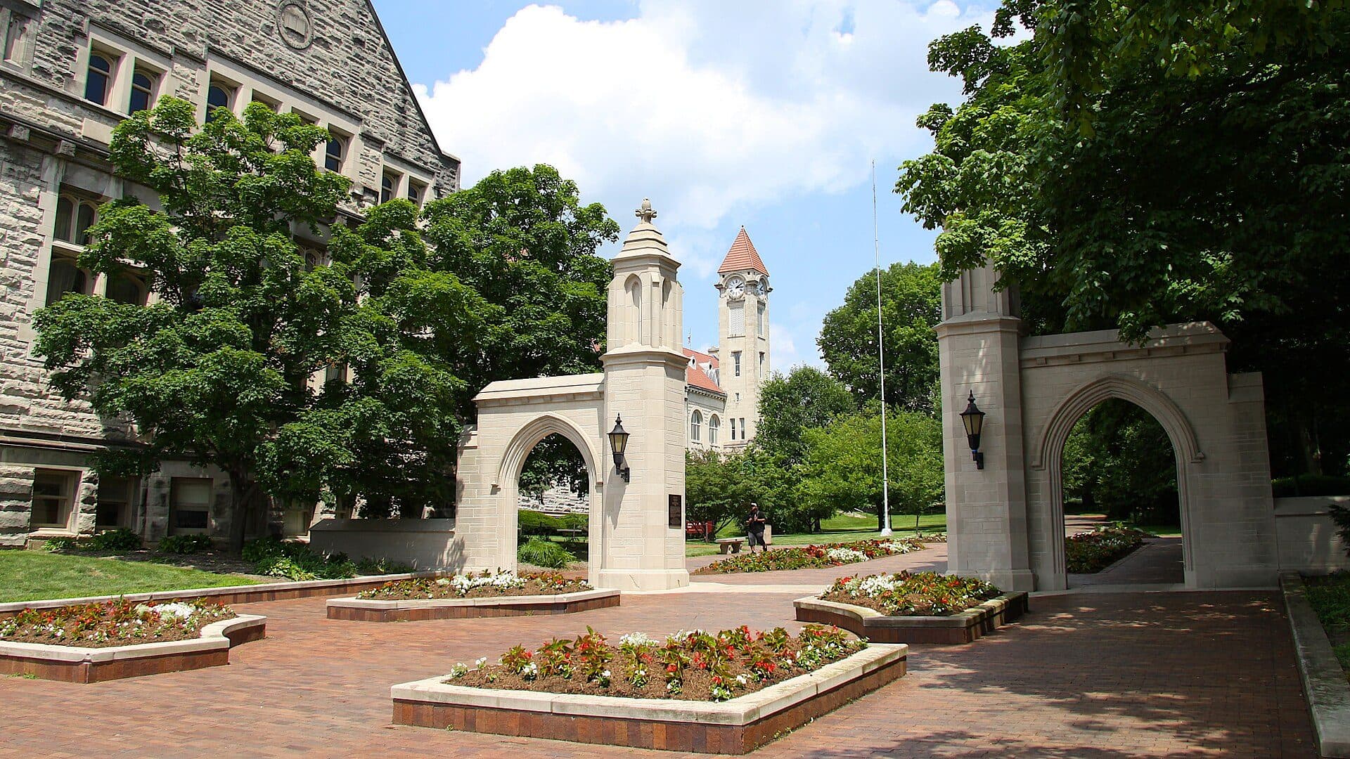 The limestone Sample Gates at Indiana University Bloomington, framed by leafy trees and flower beds, with a clock tower rising behind them under a blue sky