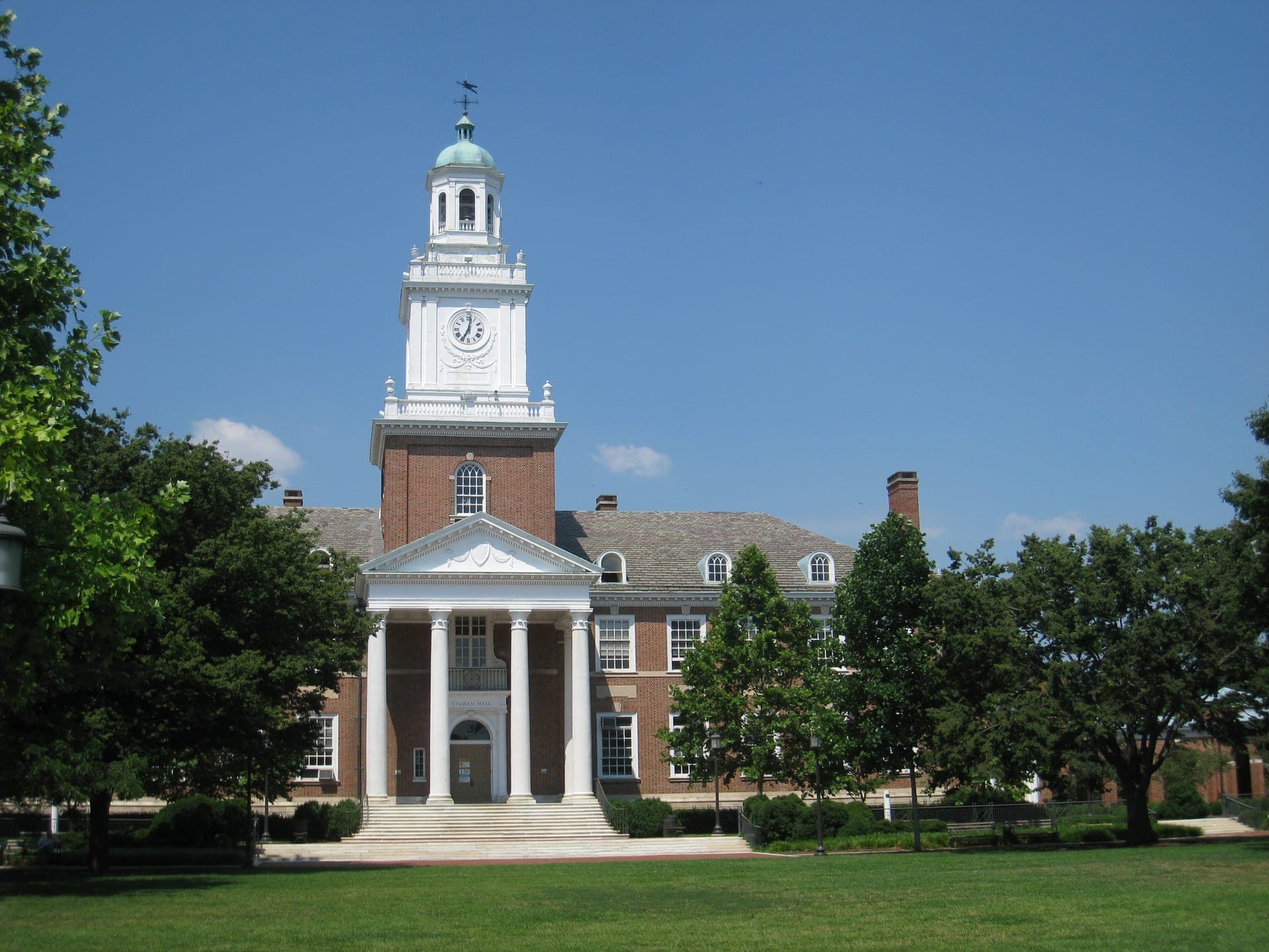Gilman Hall at Johns Hopkins, a red-brick building with a white columned portico and clock tower, behind trees and a lawn