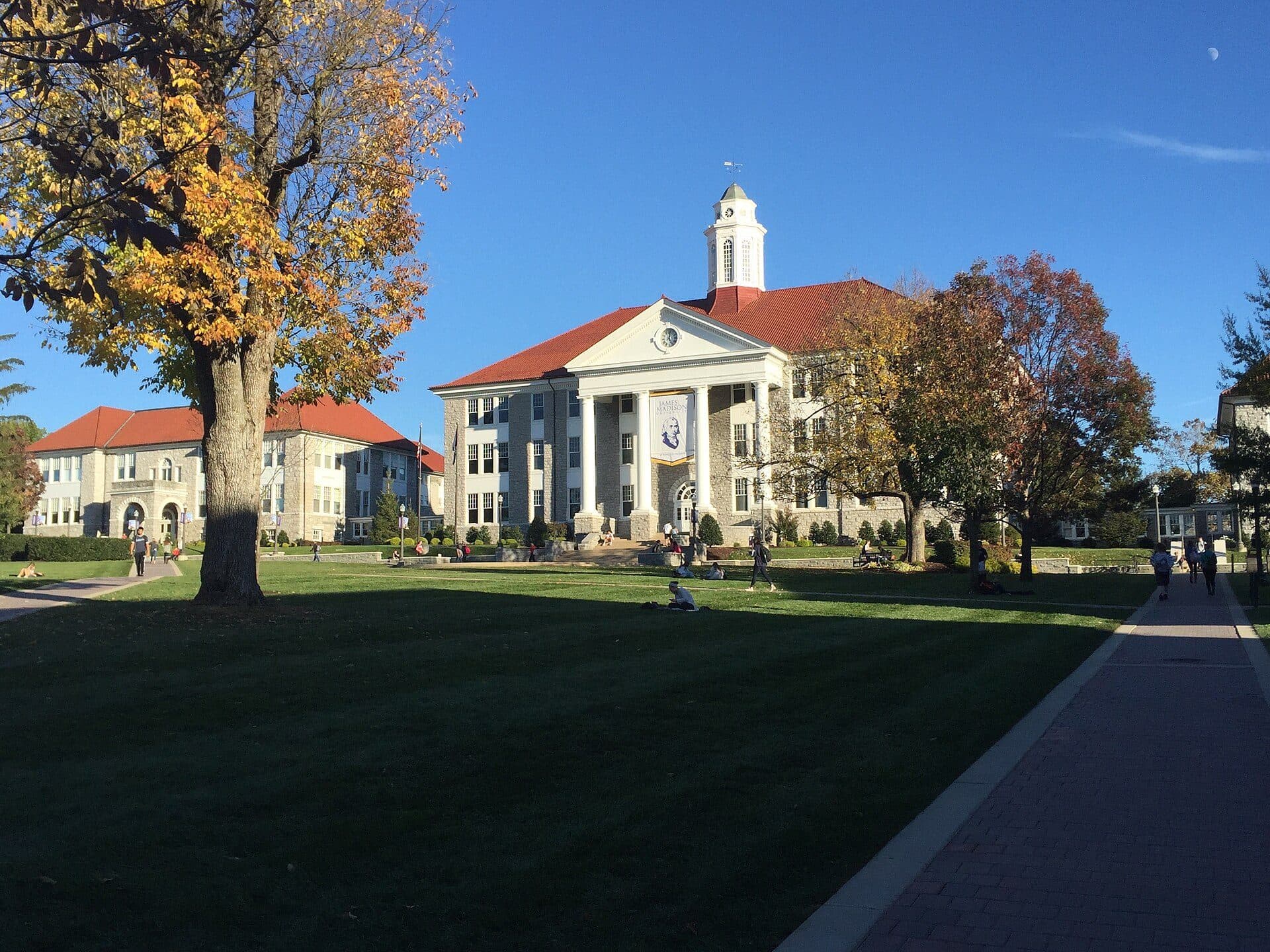 Wilson Hall, a columned stone building with a white cupola and red roof, at the end of a wide lawn with students and early fall trees under a blue sky