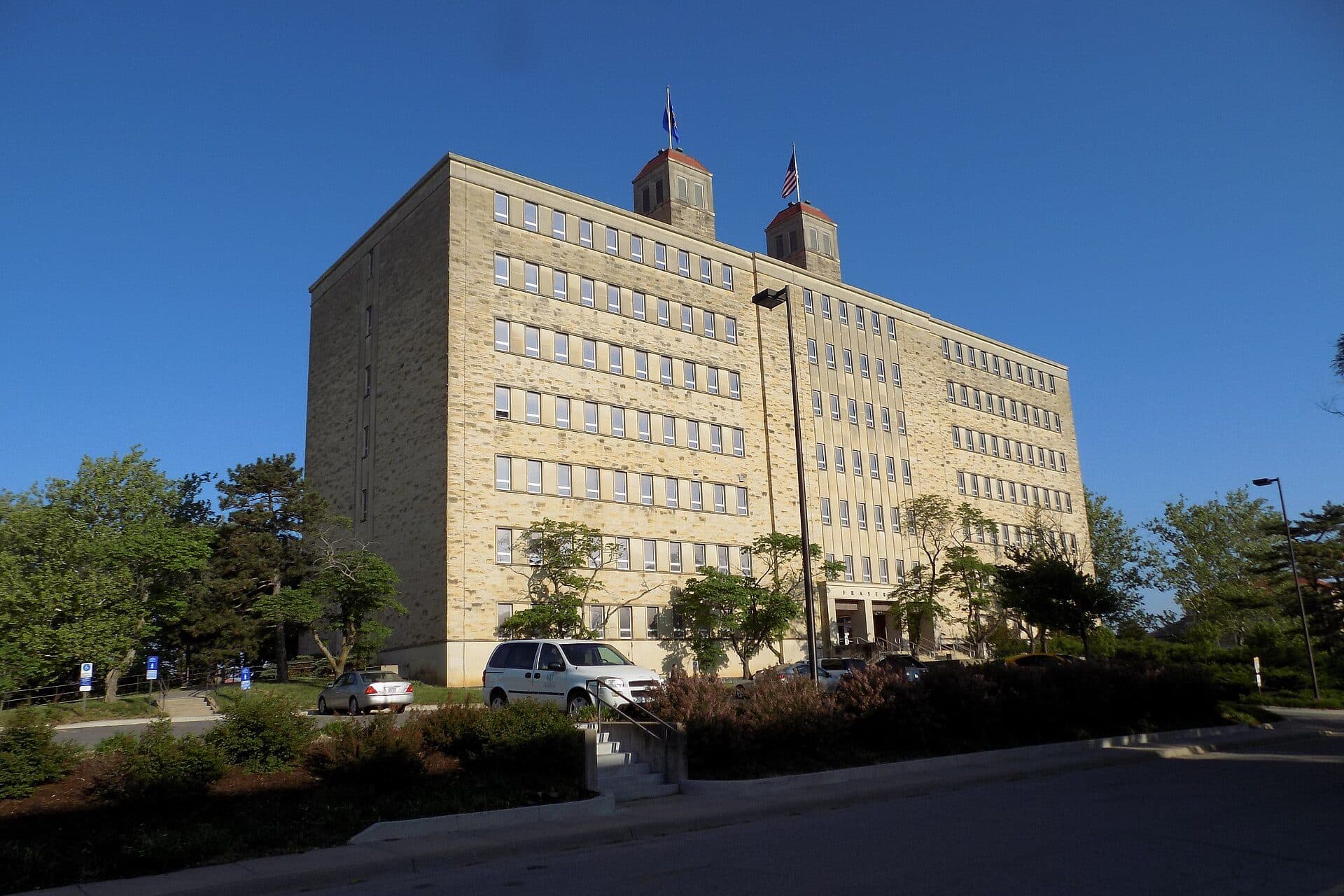 Fraser Hall, a tall limestone building with twin red-roofed towers and flags, on the University of Kansas campus under a deep blue sky