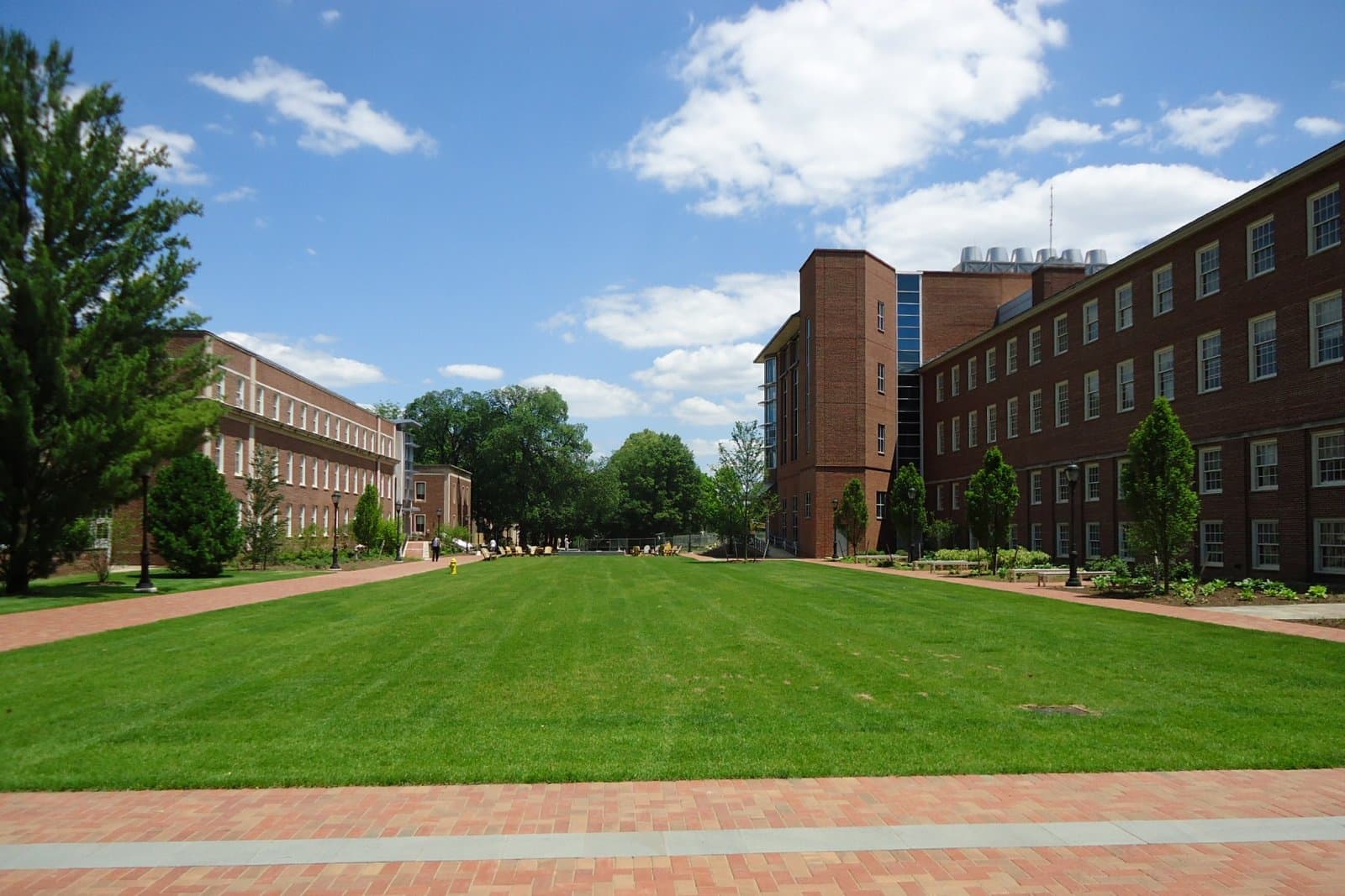 A wide mowed lawn framed by red-brick academic buildings and a glass-fronted science building under a blue sky with white clouds, with a brick walkway in the foreground