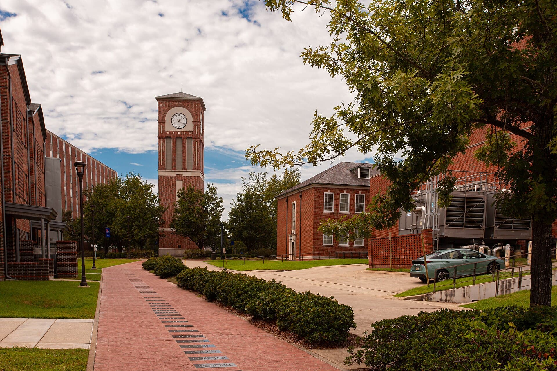Alumni Walk at Louisiana Tech, a wide brick-paved walkway lined with trees and lampposts leading west toward a brick clock tower, with Aswell Suites on the left and Howard Auditorium on the right