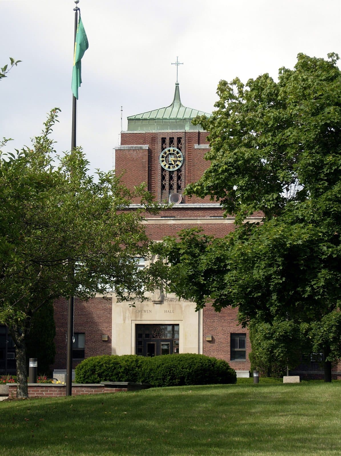 The red-brick Grewen Hall clock tower with a green copper roof and cross, framed by leafy trees and a flagpole above a green lawn at Le Moyne College