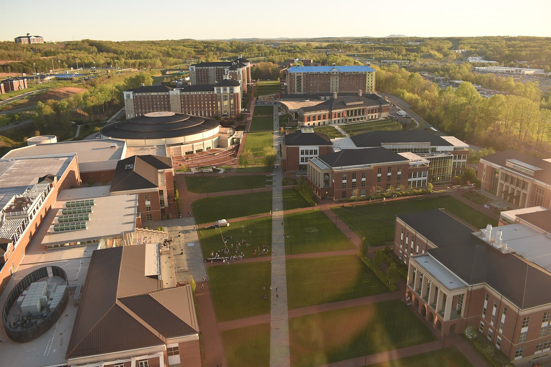 Aerial view of red-brick academic buildings, a round domed arena and long green lawns crossed by walkways on the Liberty University campus at sunset, with wooded hills beyond