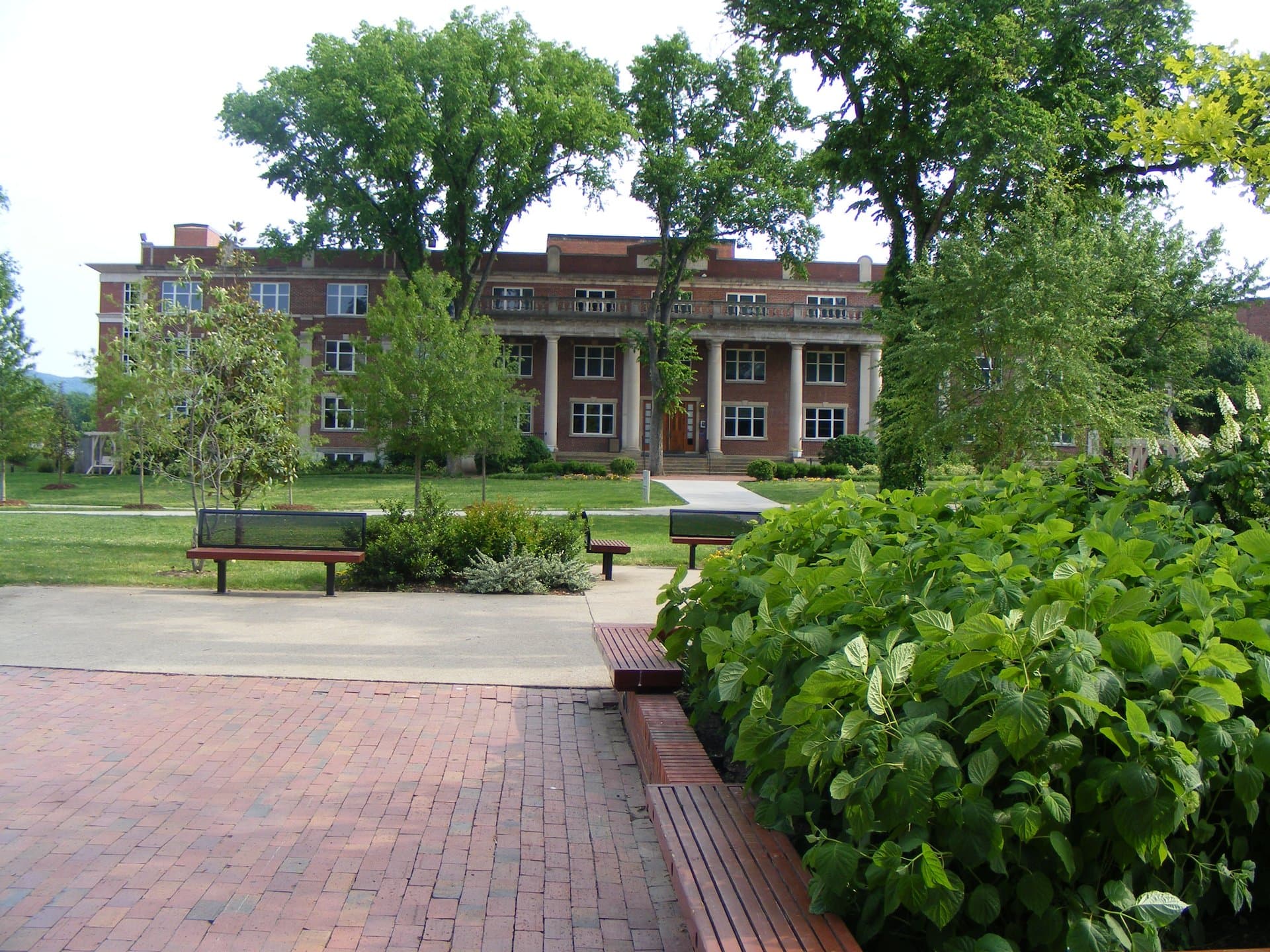 A red-brick building with white columns across its front at Lipscomb University in Nashville, seen across a green lawn with shade trees, benches and a brick walkway lined with leafy shrubs