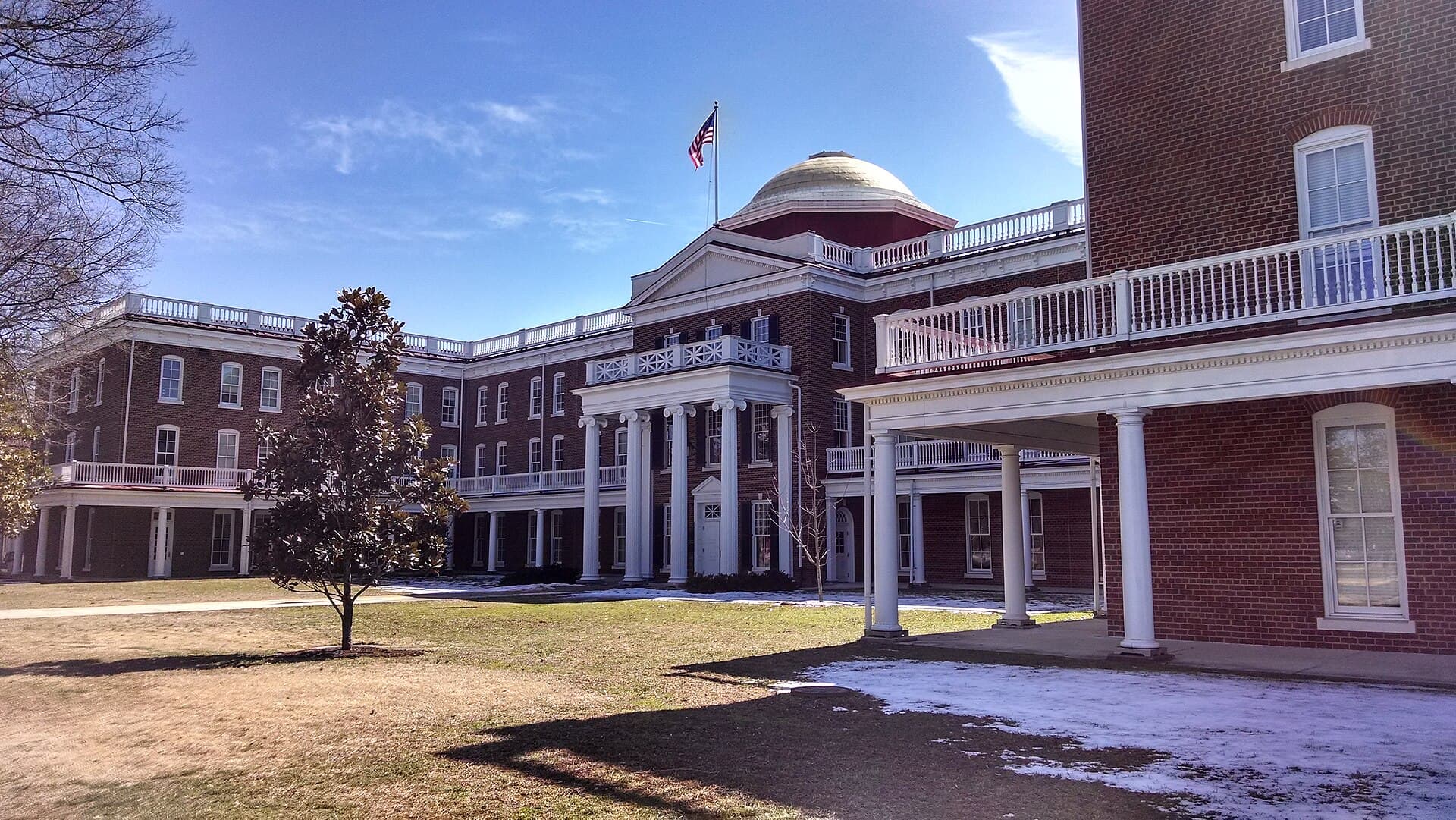 A red-brick building with white columned porches, balconies and a dome topped by an American flag at Longwood University, with a magnolia tree on the lawn