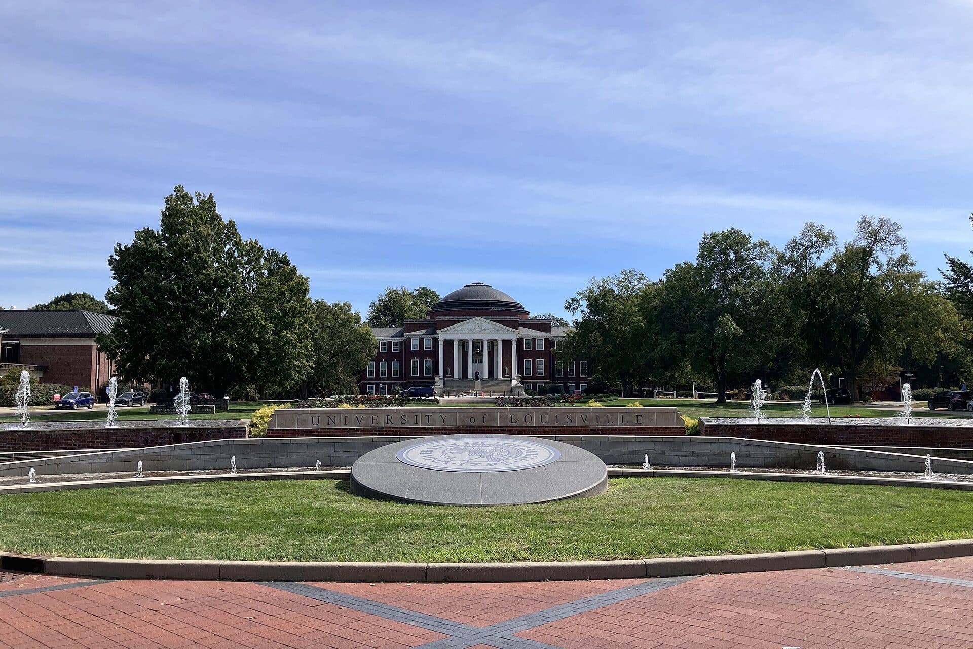 Grawemeyer Hall's white dome and columned portico at the end of a wide lawn and plaza on UofL's Belknap Campus, framed by trees