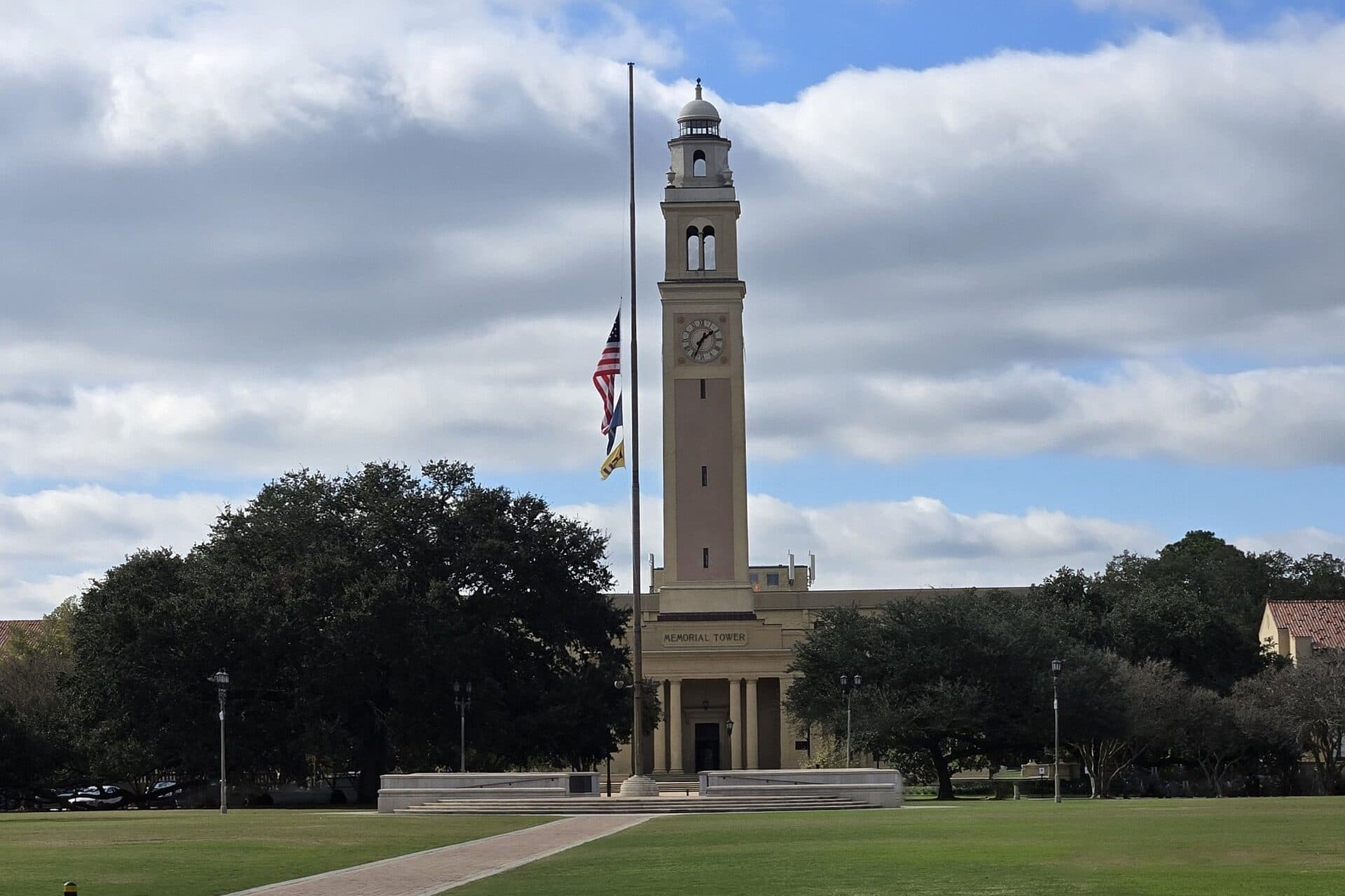 LSU's tall cream-colored Memorial Tower with a clock face rising between dark live oaks across the wide green Parade Ground under a cloudy sky