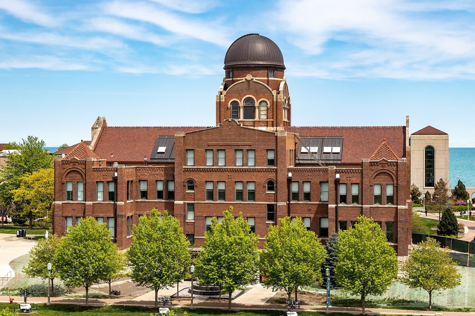 Cudahy Science Hall's red-brick facade and copper observatory dome on Loyola's Lake Shore Campus, with young trees in front and Lake Michigan visible at the edge