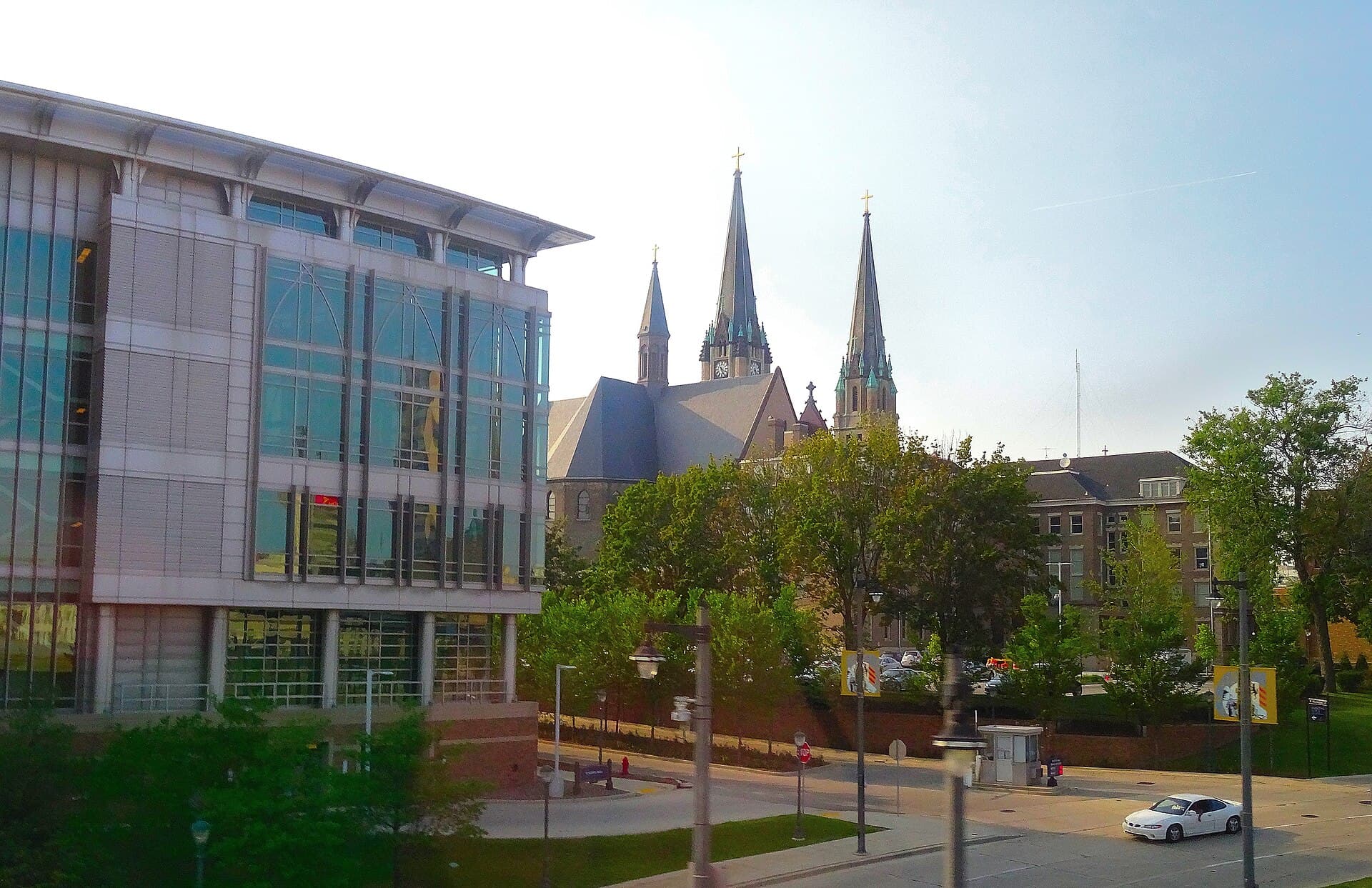 Marquette's campus on a summer evening, with a modern glass-and-stone building on the left, the tall twin Gothic spires of a stone church rising behind green trees in the center and a red-brick building on the right, above a quiet street