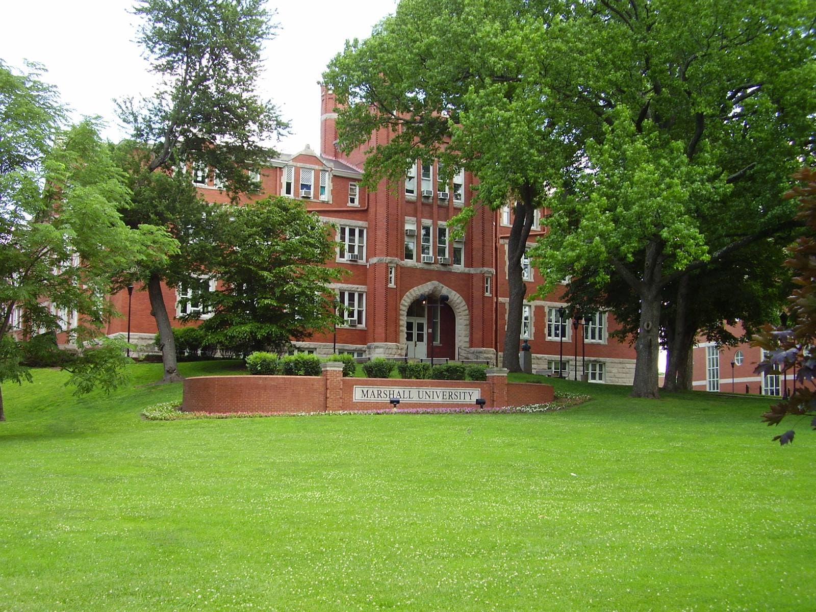 A red-brick Victorian building with a central tower, a stone-arched entrance and rows of white-trimmed windows, partly framed by tall leafy trees, behind a wide green lawn and a low brick sign reading Marshall University