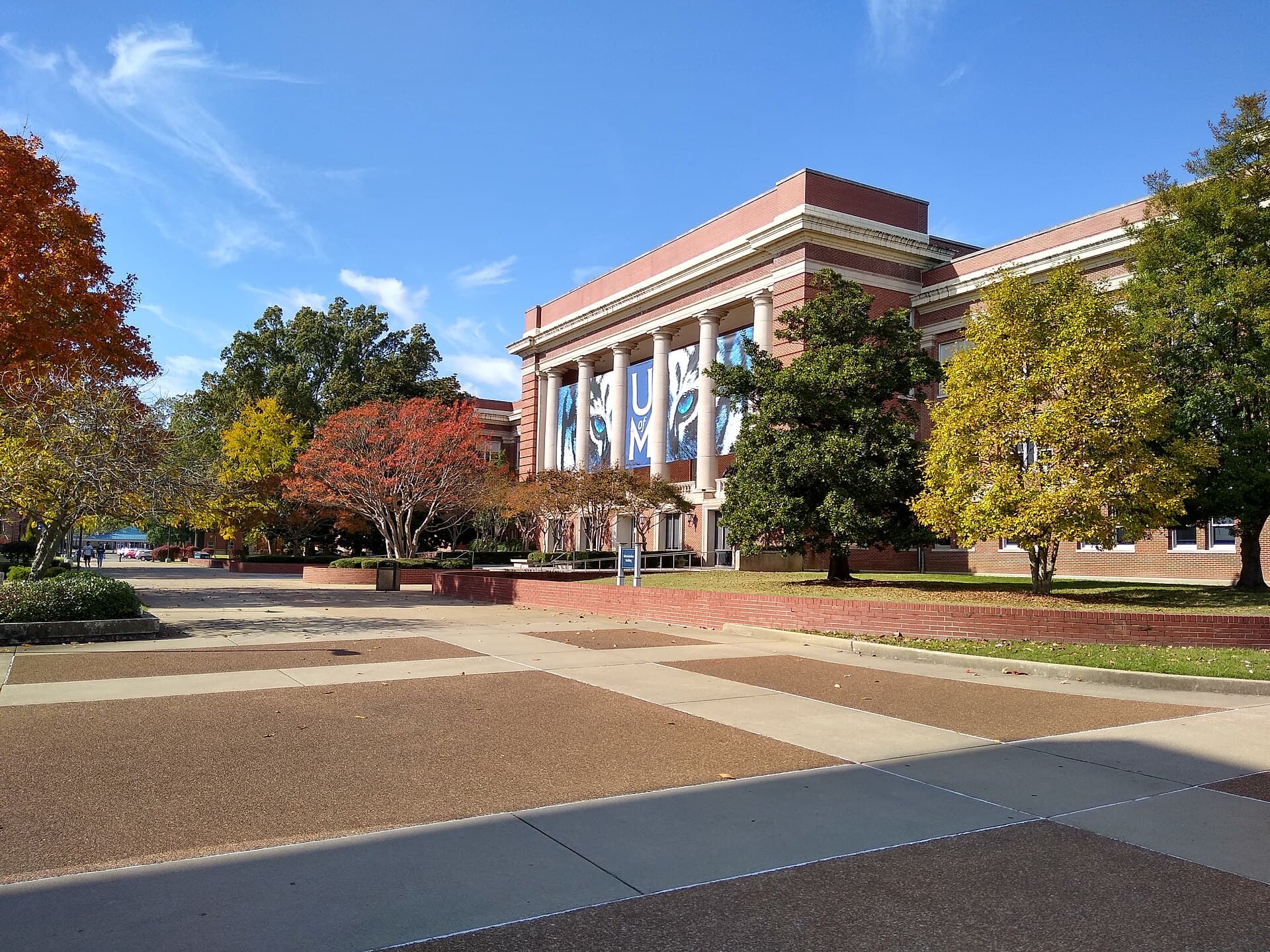 The University of Memphis Administration Building, a red-brick building with a white-columned portico and a tiger-eye banner, framed by red and yellow fall trees beside a wide paved walk